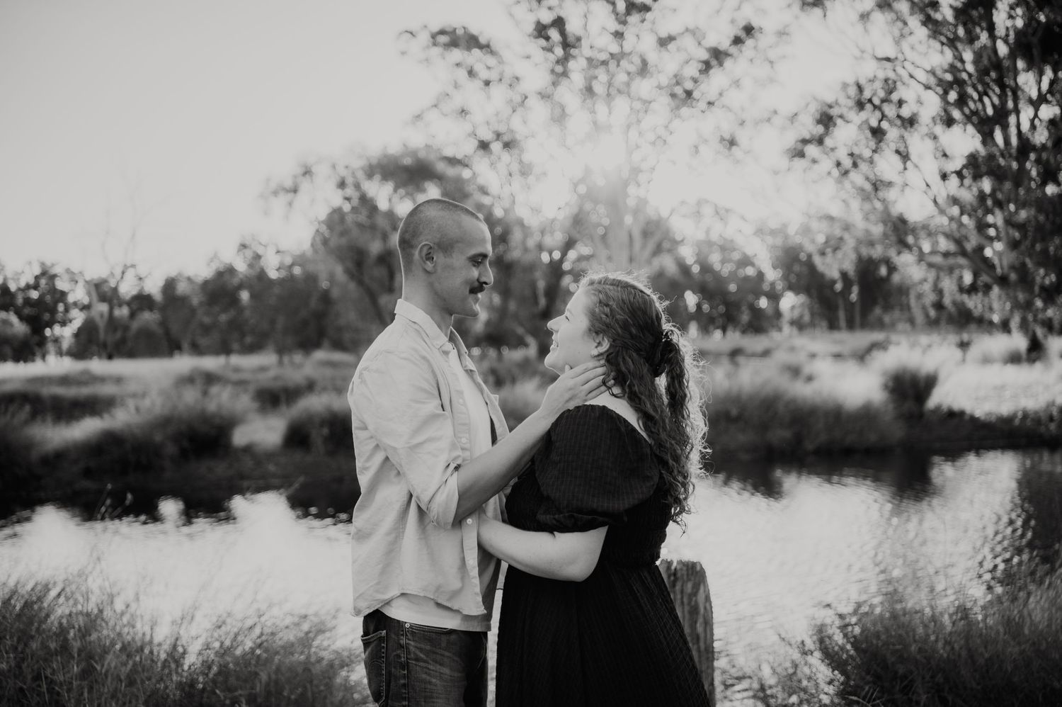 Black and white photo of a couple sharing a romantic moment outdoors near a pond surrounded by trees.