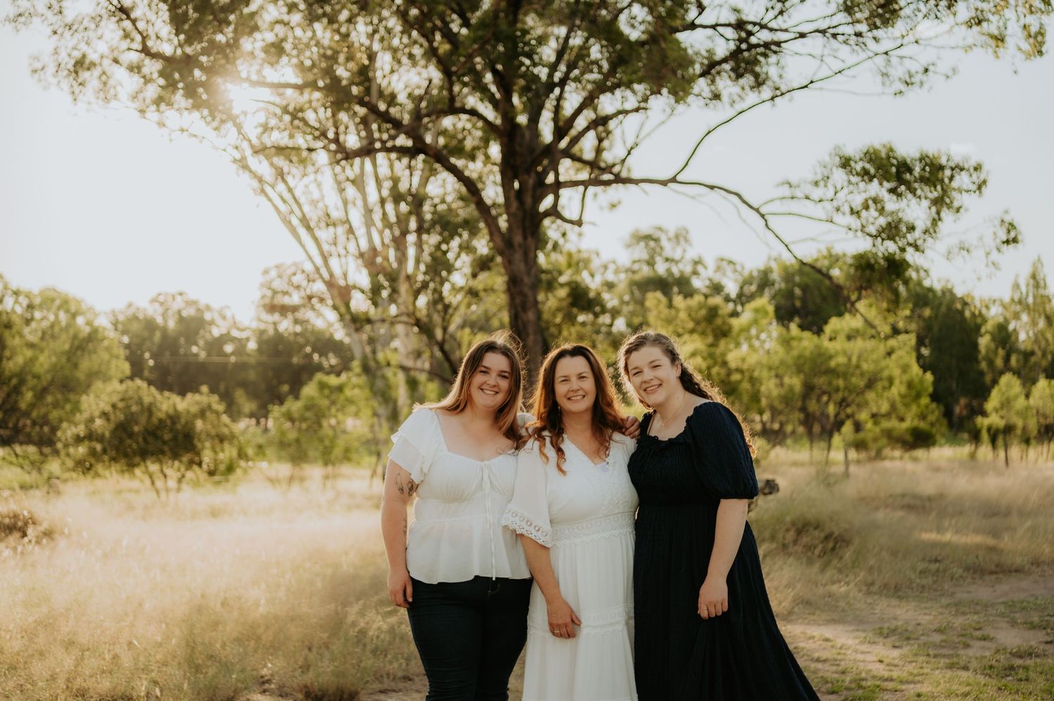 Three women smiling together outdoors in golden hour light, surrounded by trees in a natural field setting.