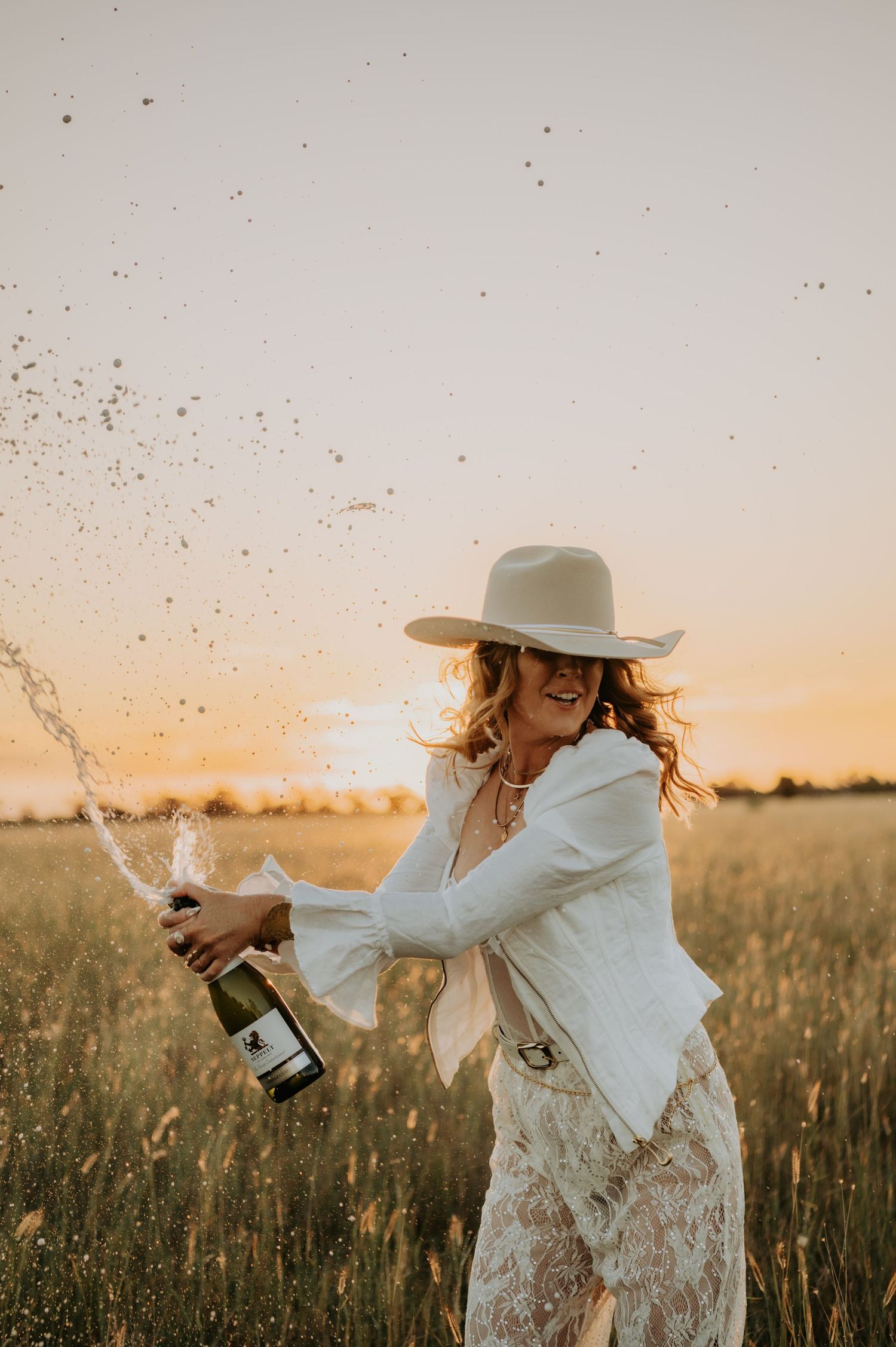 Person in white dress spraying champagne in sunset field.