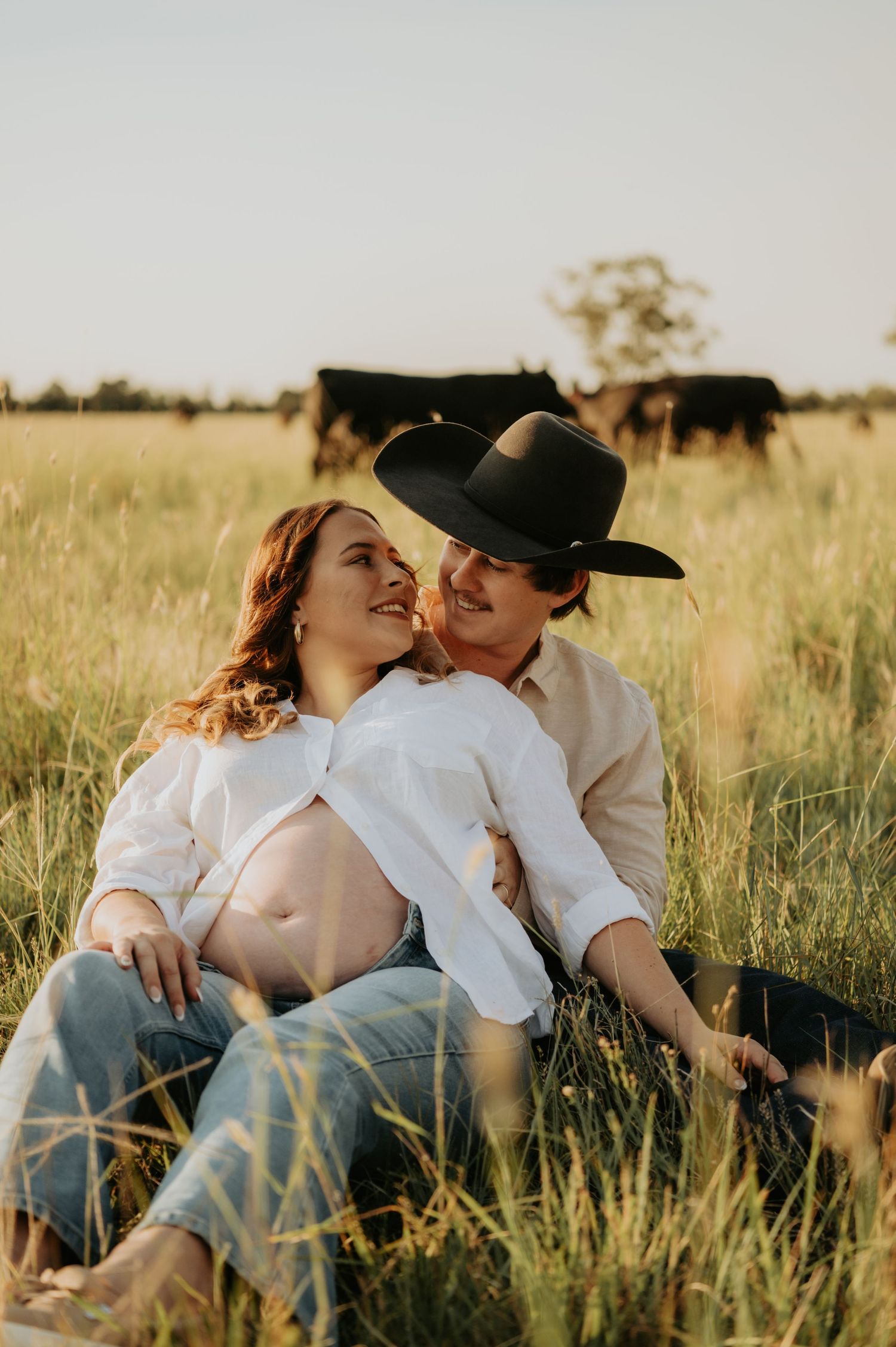 A romantic couple lounges together in a golden wheat field during sunset, creating an intimate countryside moment.