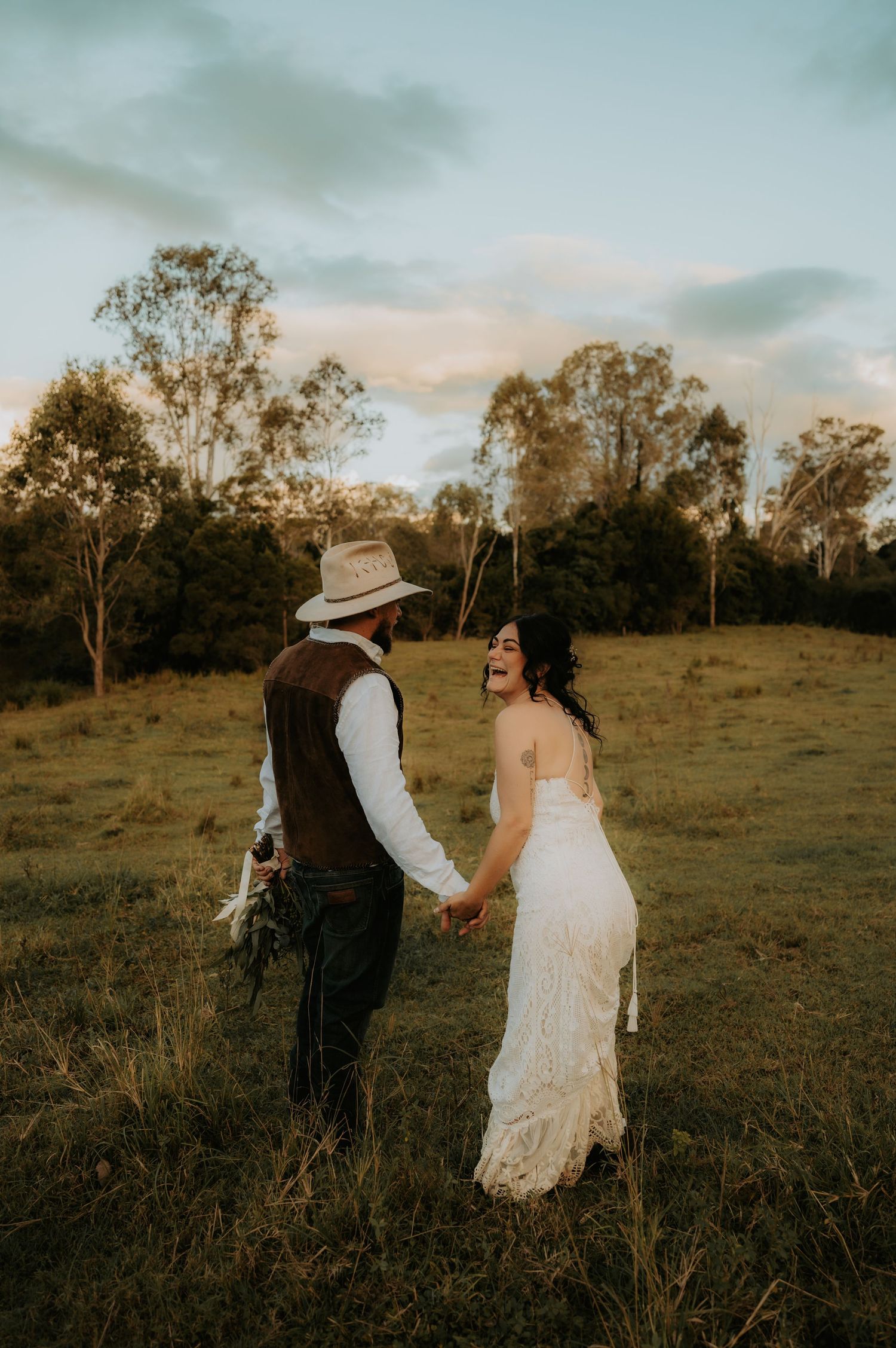 A bride in lace gown and groom in cowboy hat hold hands laughing in a golden sunlit field at sunset.