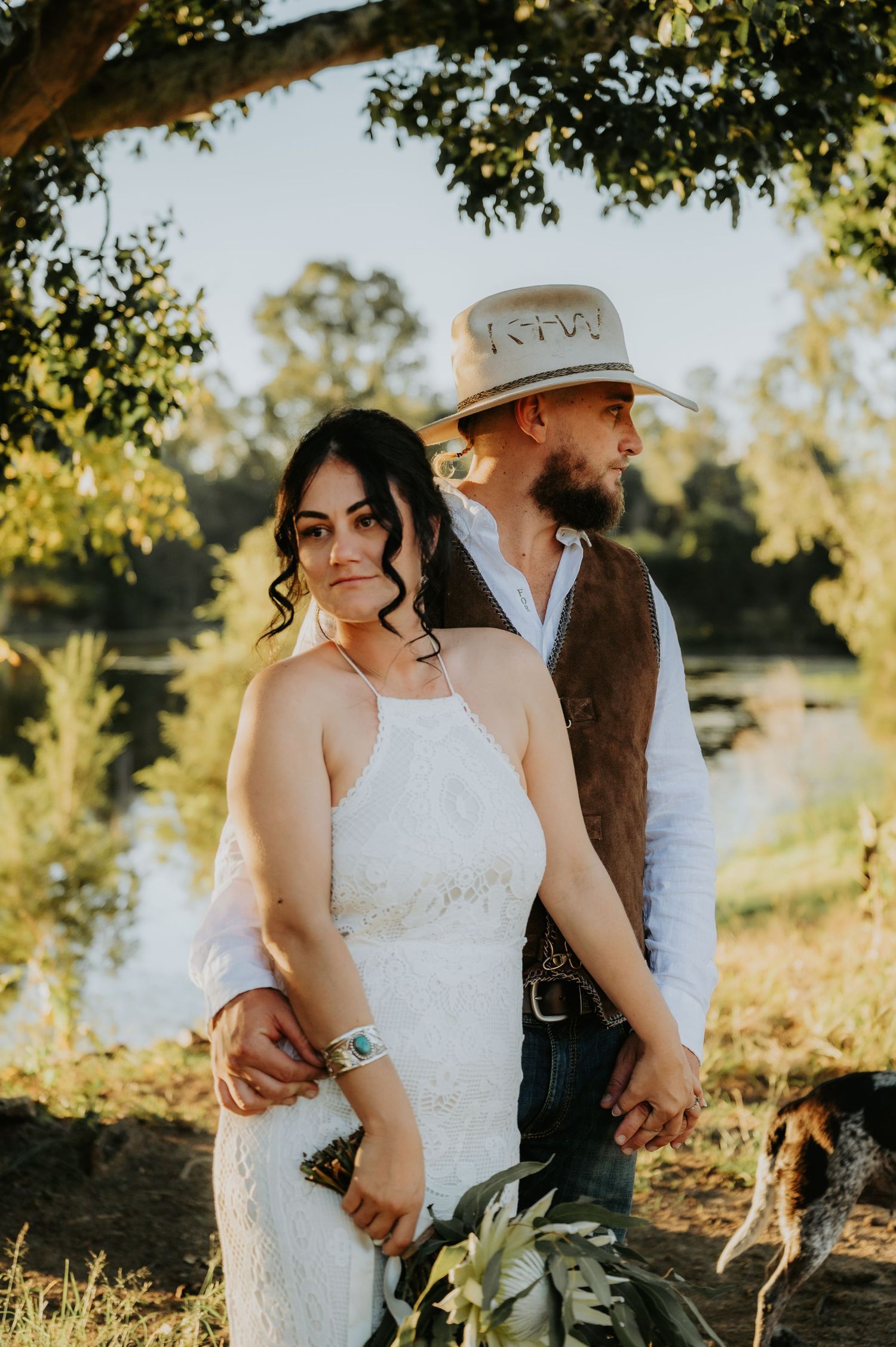 Bride in white halter gown and groom in cowboy hat and vest hold hands outdoors near a sunlit pond.