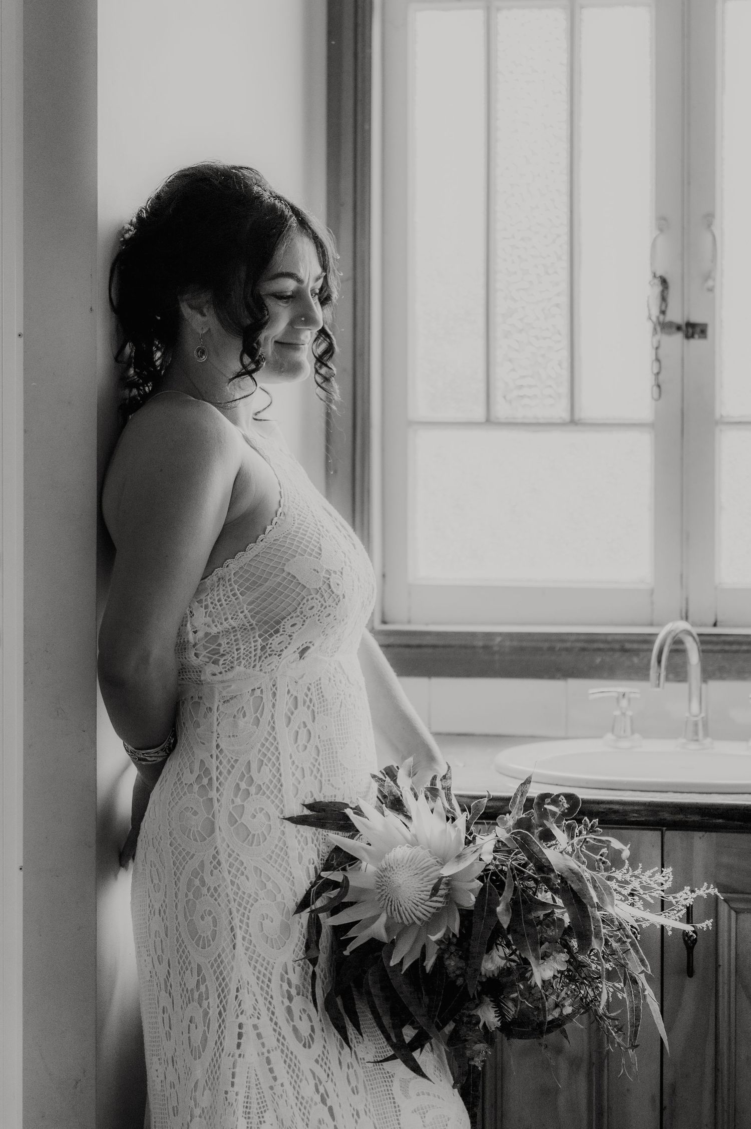 Bride in lace wedding gown holding bouquet gazes out window in black and white portrait photo.