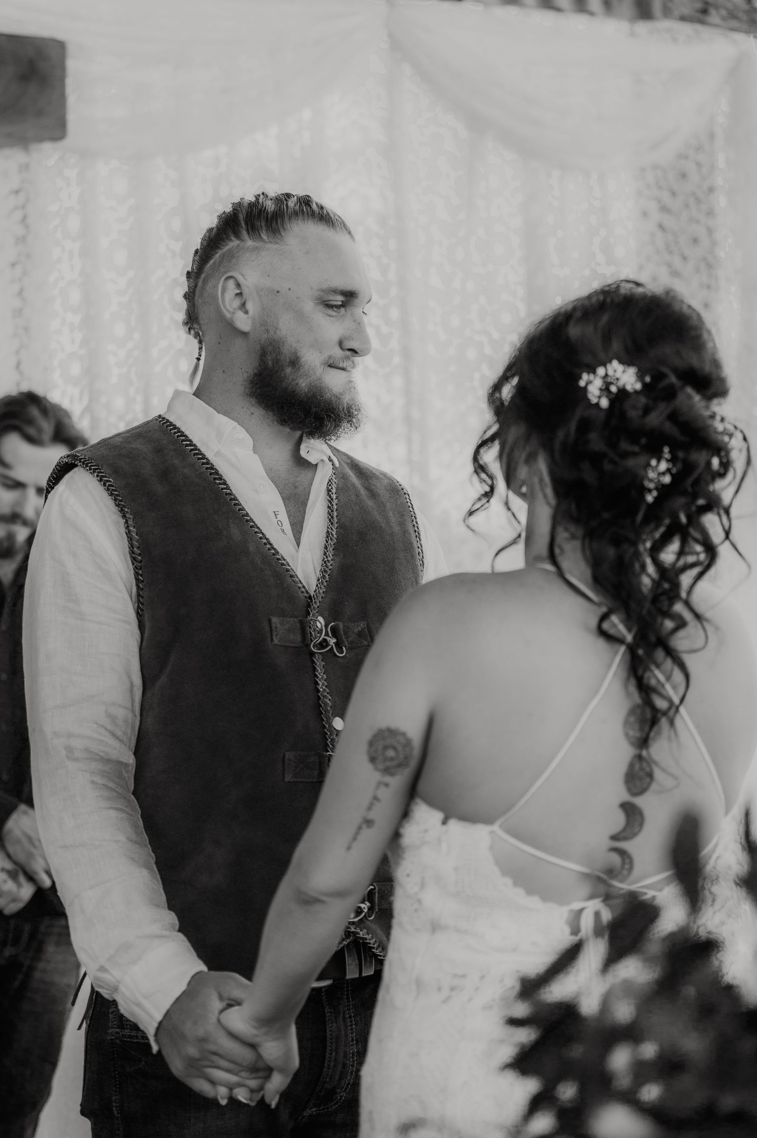 Black and white wedding photo of bearded groom holding hands with tattooed bride in lace dress during ceremony.