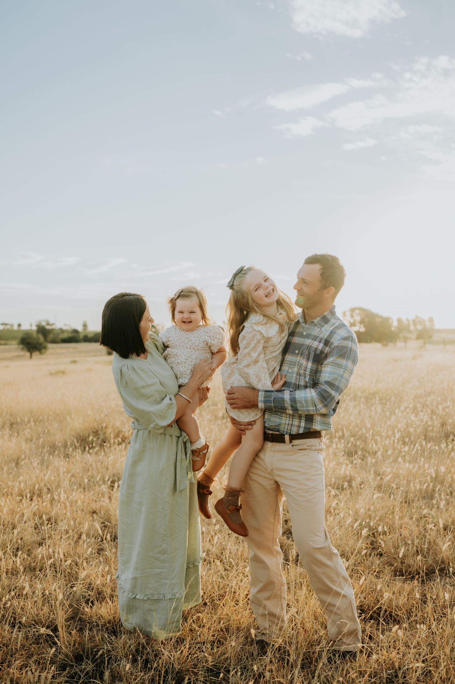 Family of four laughing together in a golden field at sunset, parents holding two young daughters in a warm outdoor portrait.