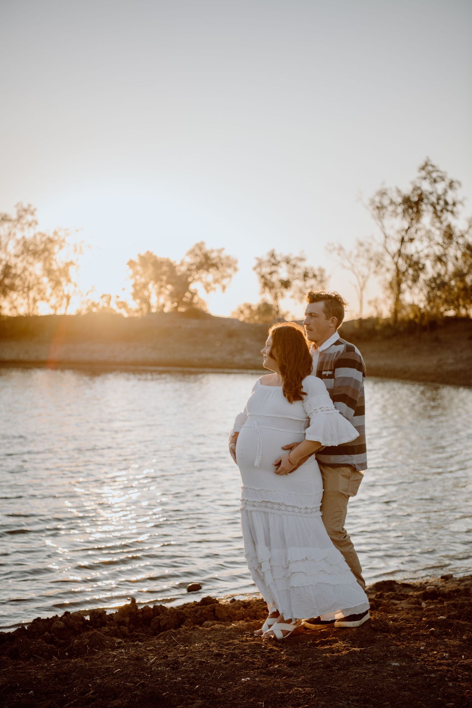 A couple embraces by a lake at sunset while standing on the shoreline in warm golden light.