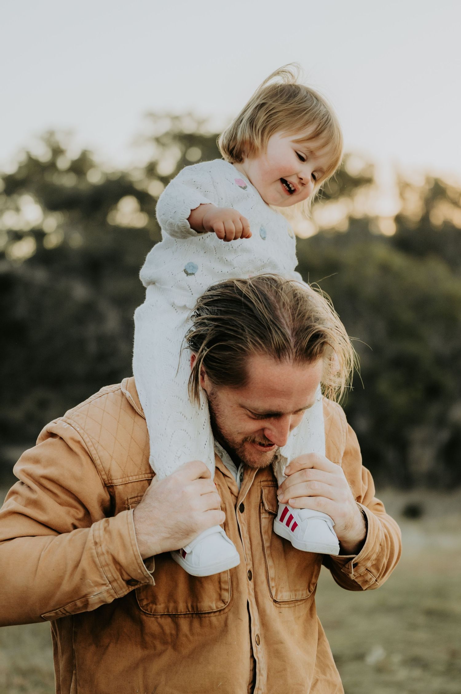 A man in a tan jacket carries a laughing toddler in white on his shoulders outdoors.