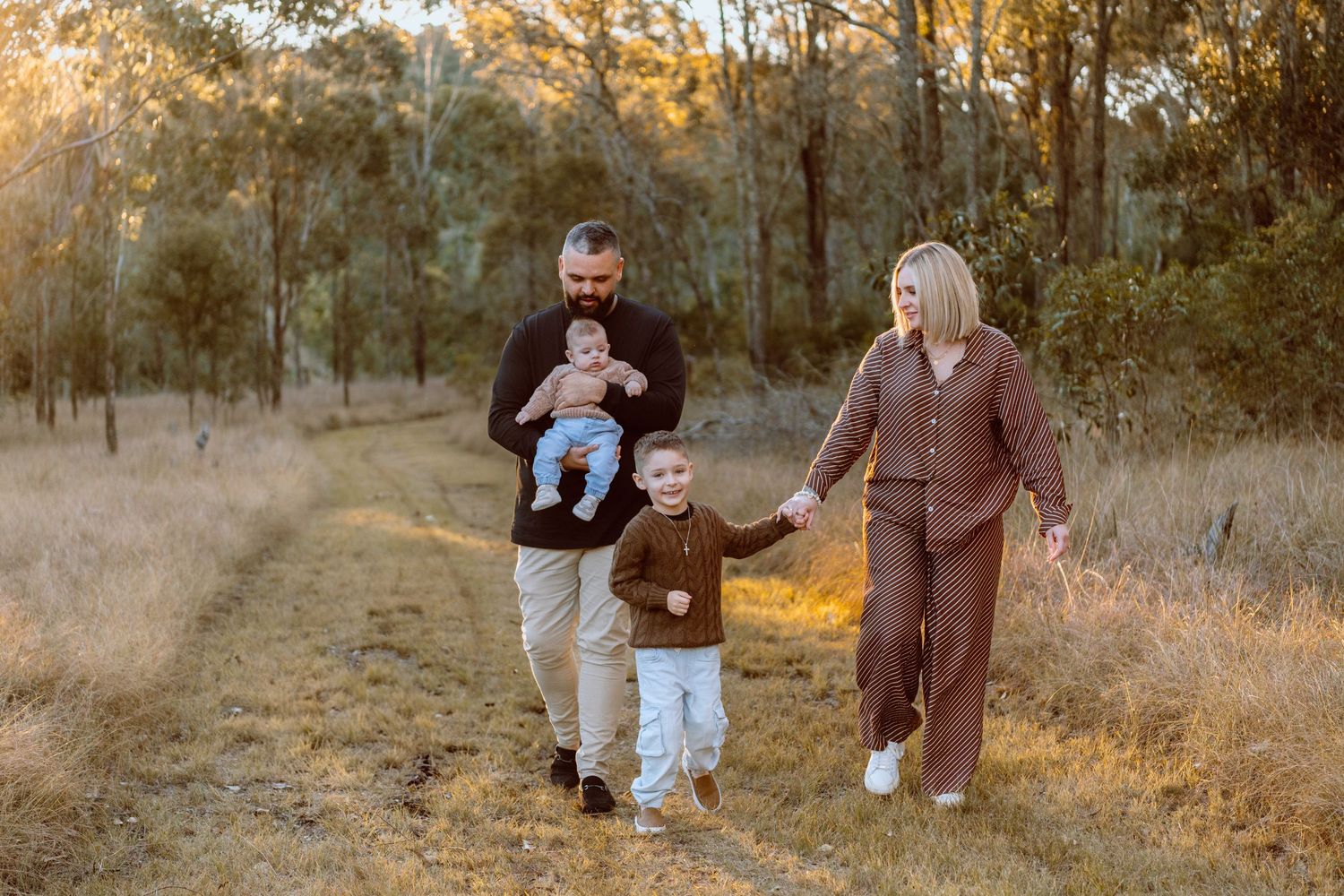 Family of four walking on a dirt path through golden Australian bushland during sunset, parents holding hands with two young children.