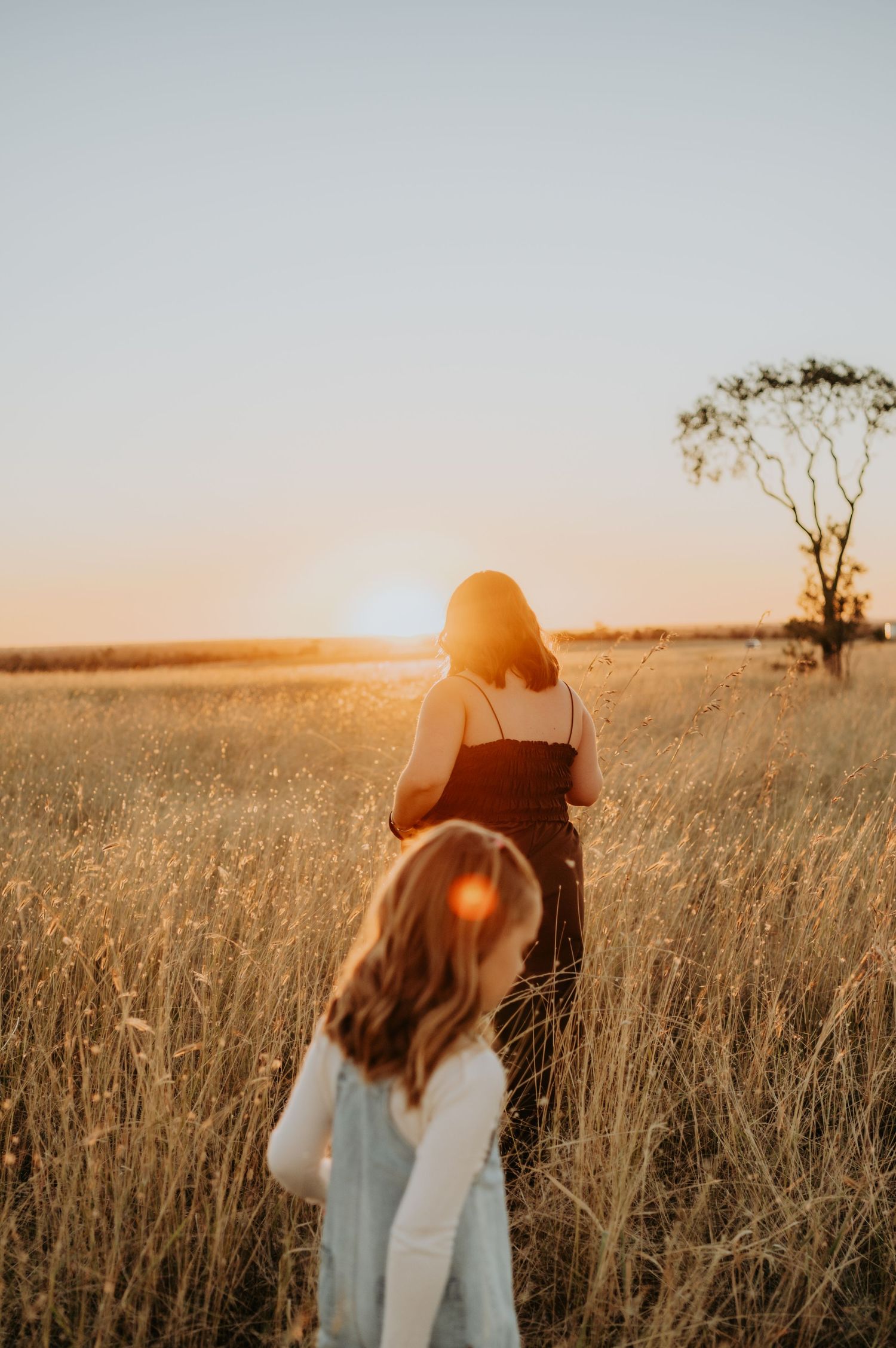 Two women walking through golden wheat fields at sunset with a lone tree on the horizon.