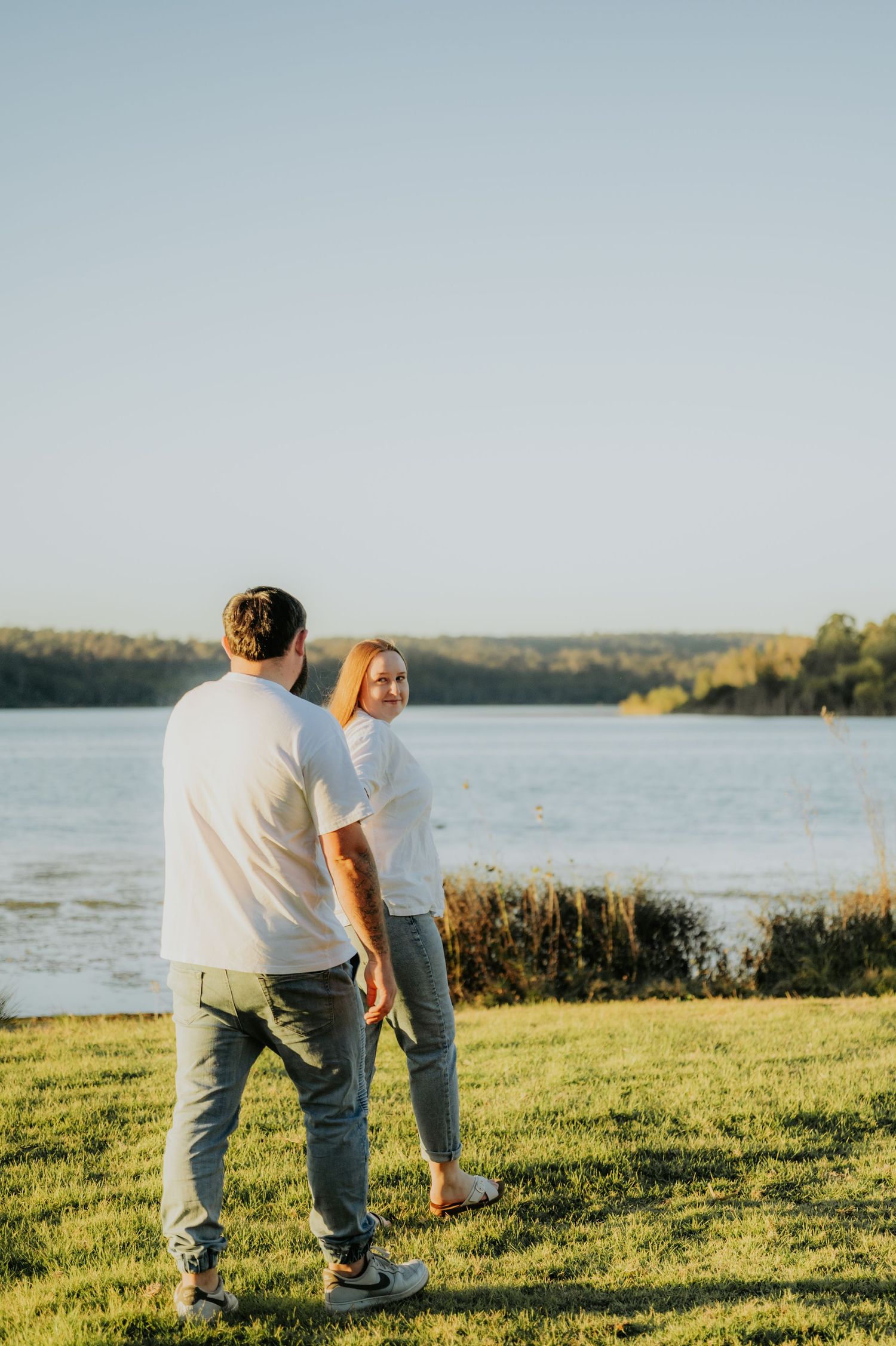 Couple holding hands by a sunlit lake, woman smiling back at camera, both wearing white shirts and jeans.