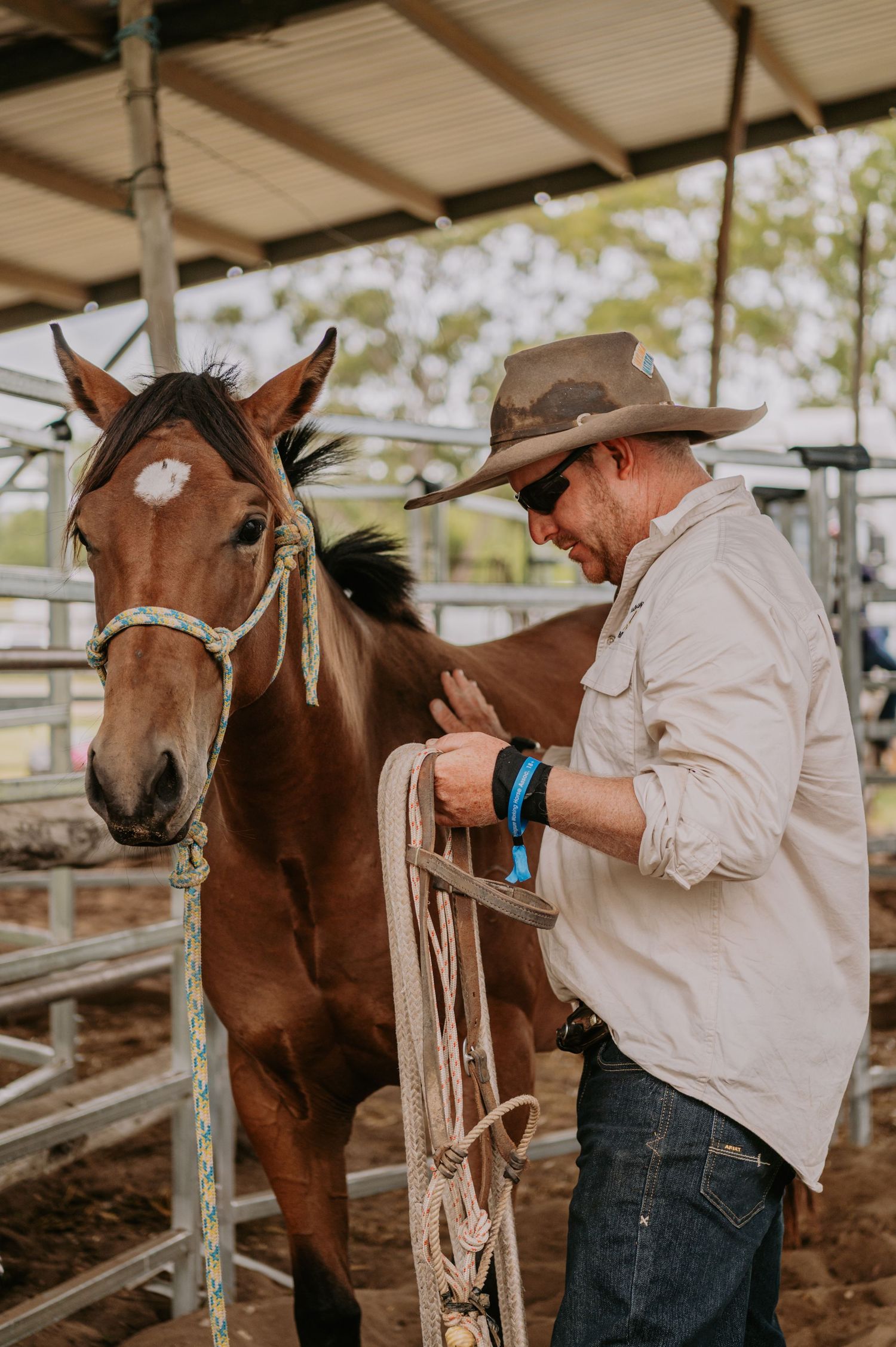 A cowboy in a wide-brim hat handles a rope lead next to a chestnut horse in a covered ranch pen.