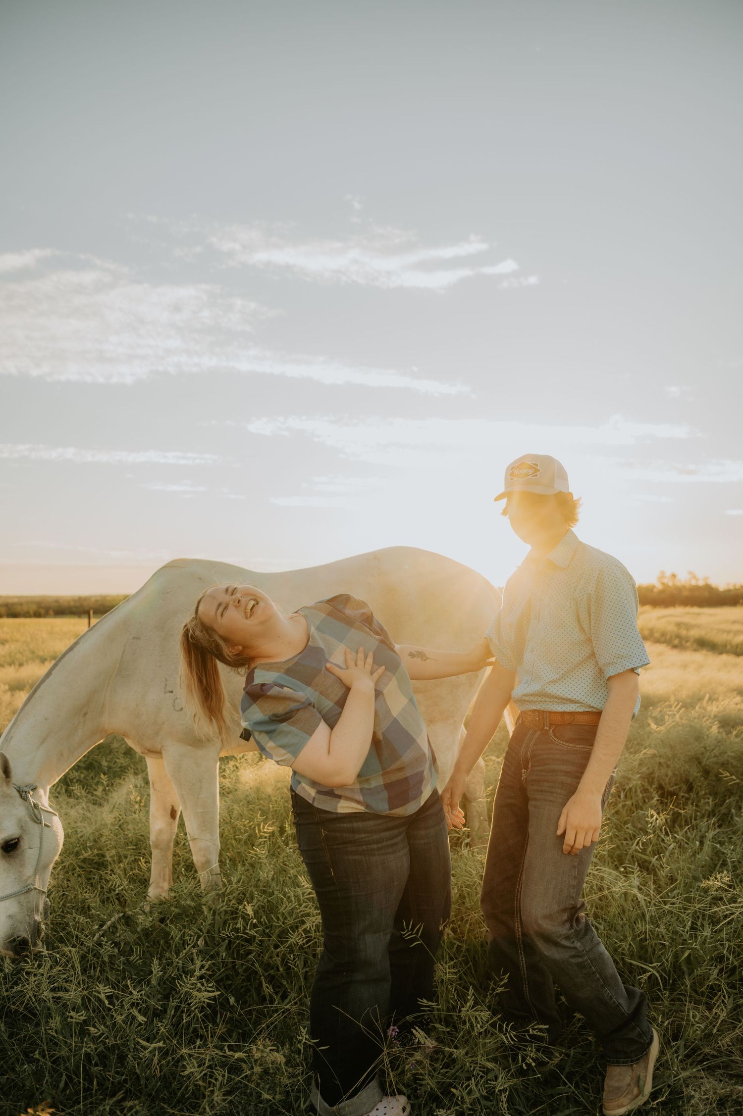 A couple interacts with a white horse in a sunlit meadow at dusk.