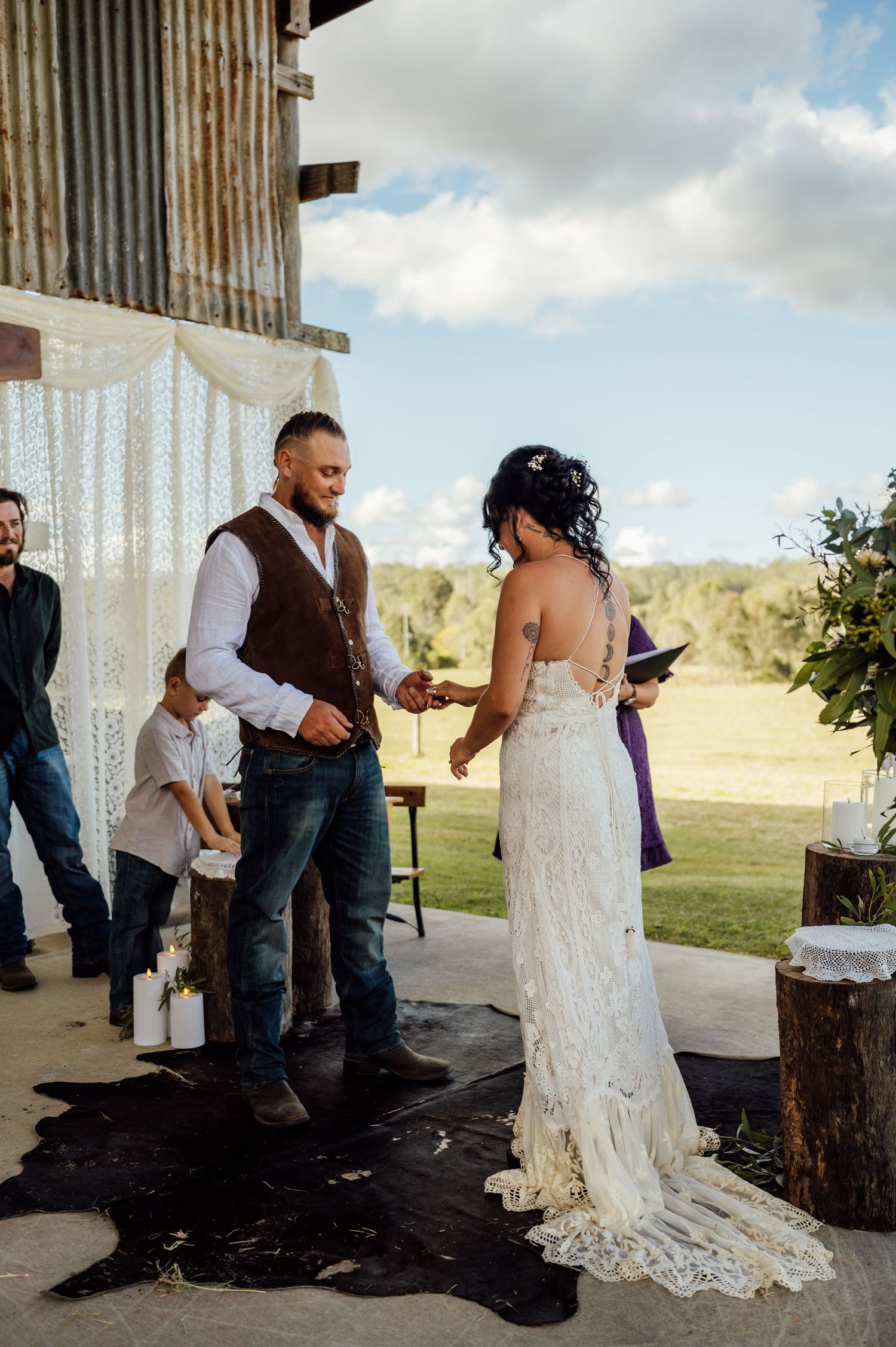 Bride and groom exchange vows at rustic barn wedding ceremony outdoors under blue sky.