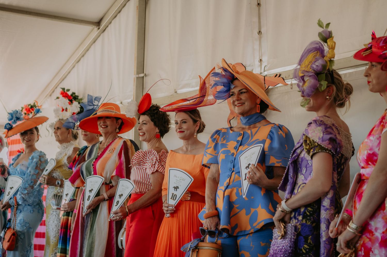 Women in colorful dresses and decorative hats pose in a row at an elegant outdoor hat competition event.