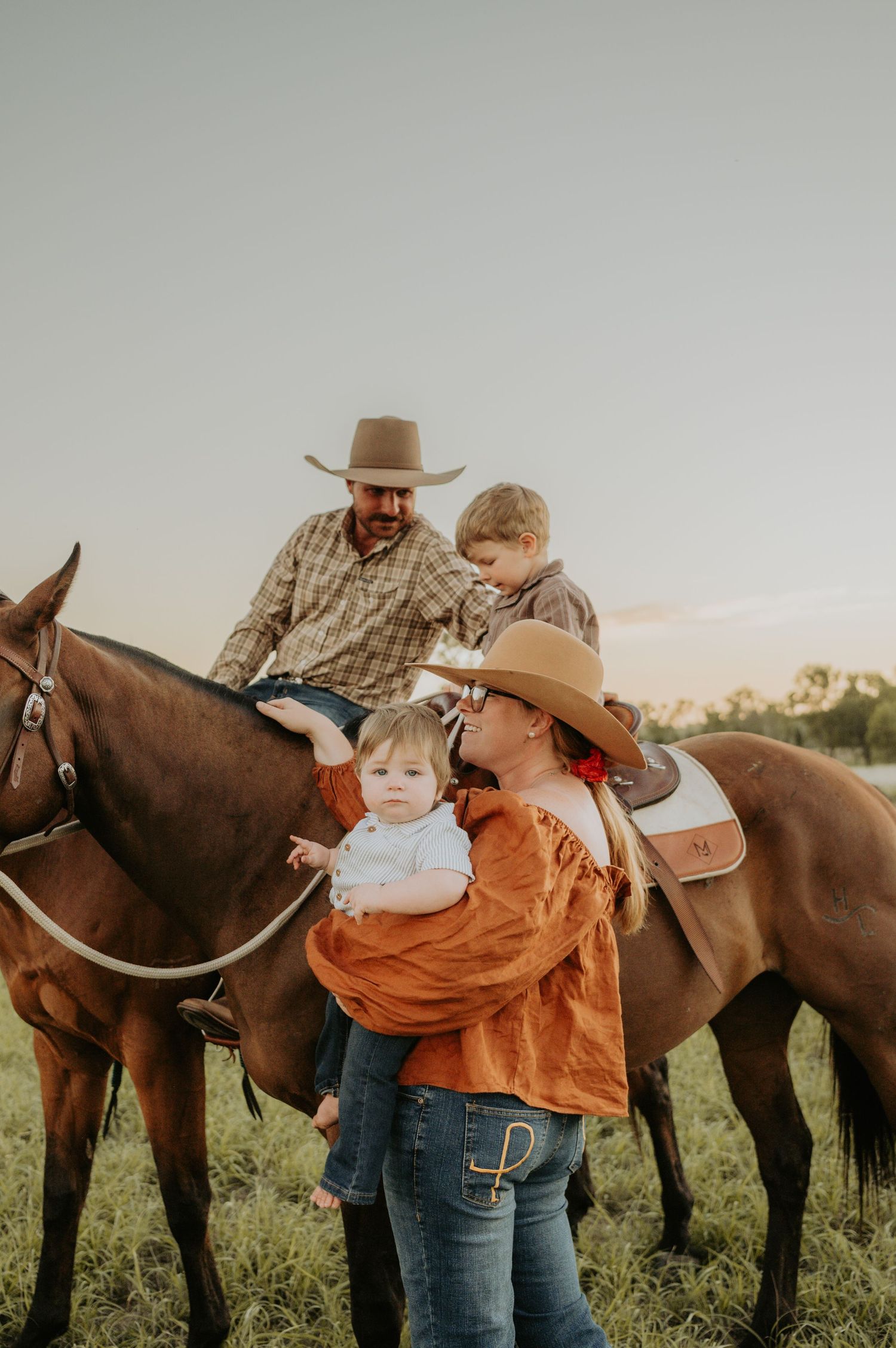 Family in cowboy hats with two horses in a field, woman holding baby while man and child sit on horseback at sunset.