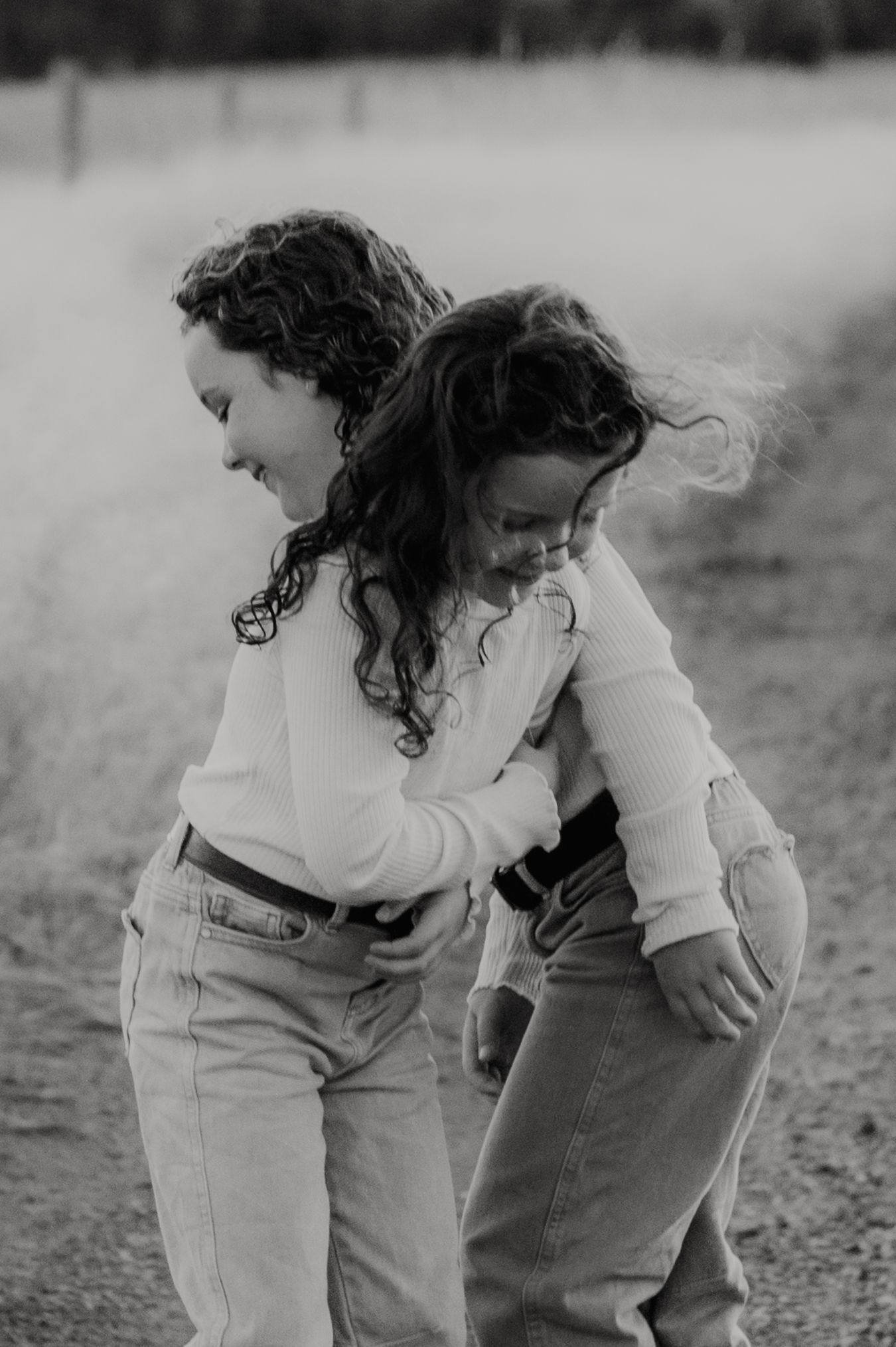 Two young girls with curly hair laugh and hug playfully on a dirt path in a black and white photo.