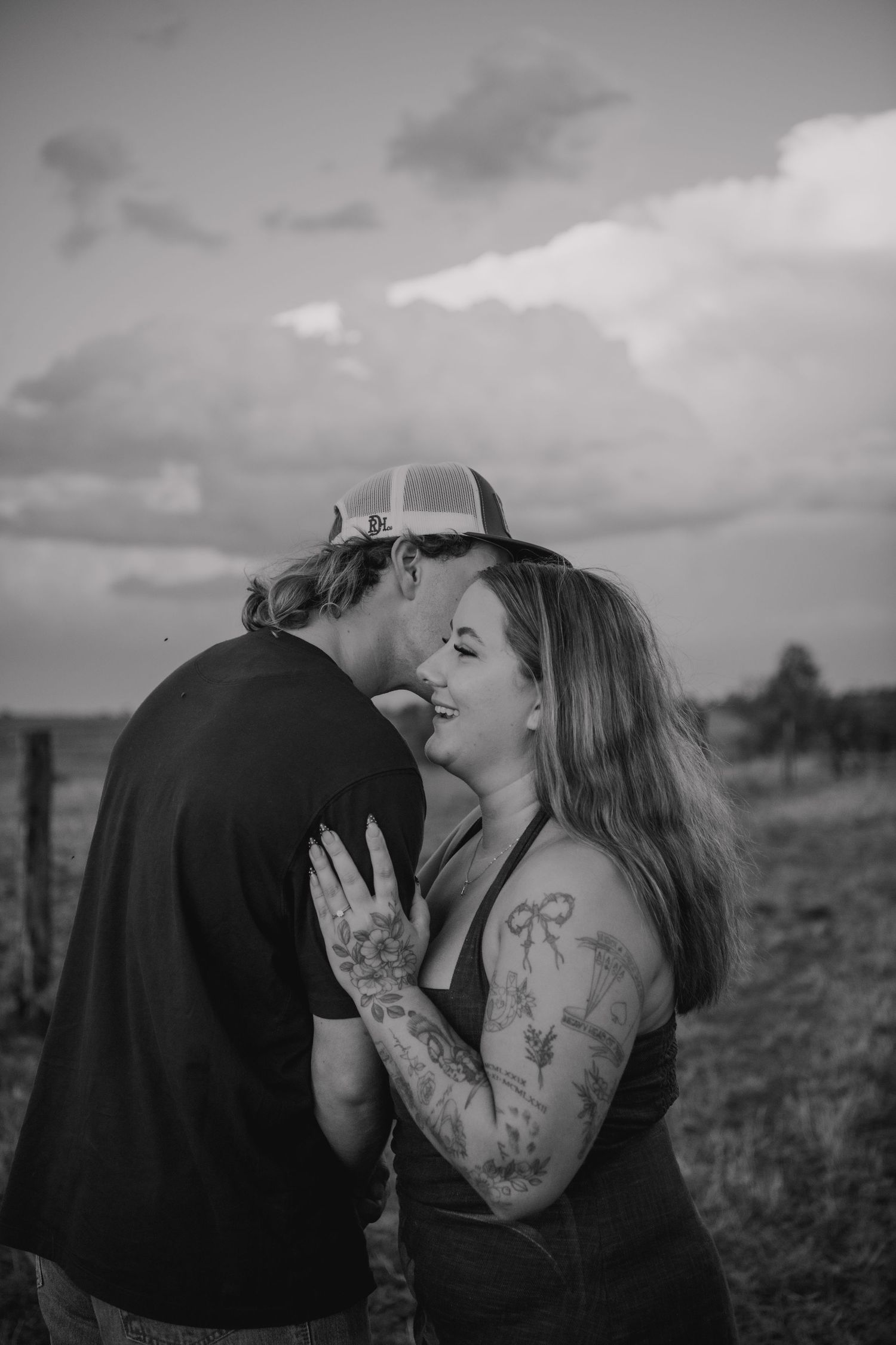 Black and white photo of a couple sharing a romantic moment outdoors, woman with tattoos smiling as man kisses her cheek.