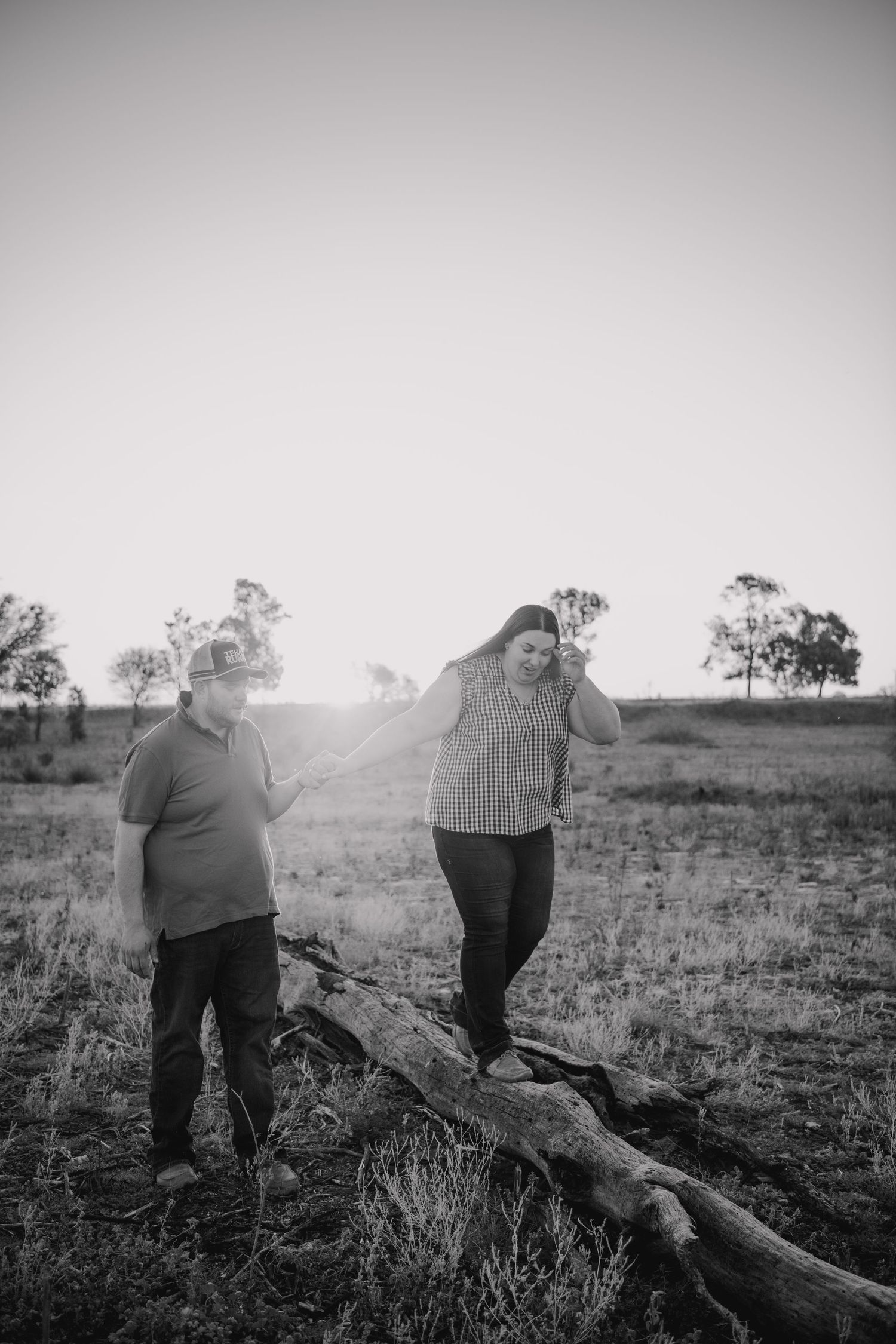 Black and white photo of a couple standing near a fallen log in an open field at sunset.