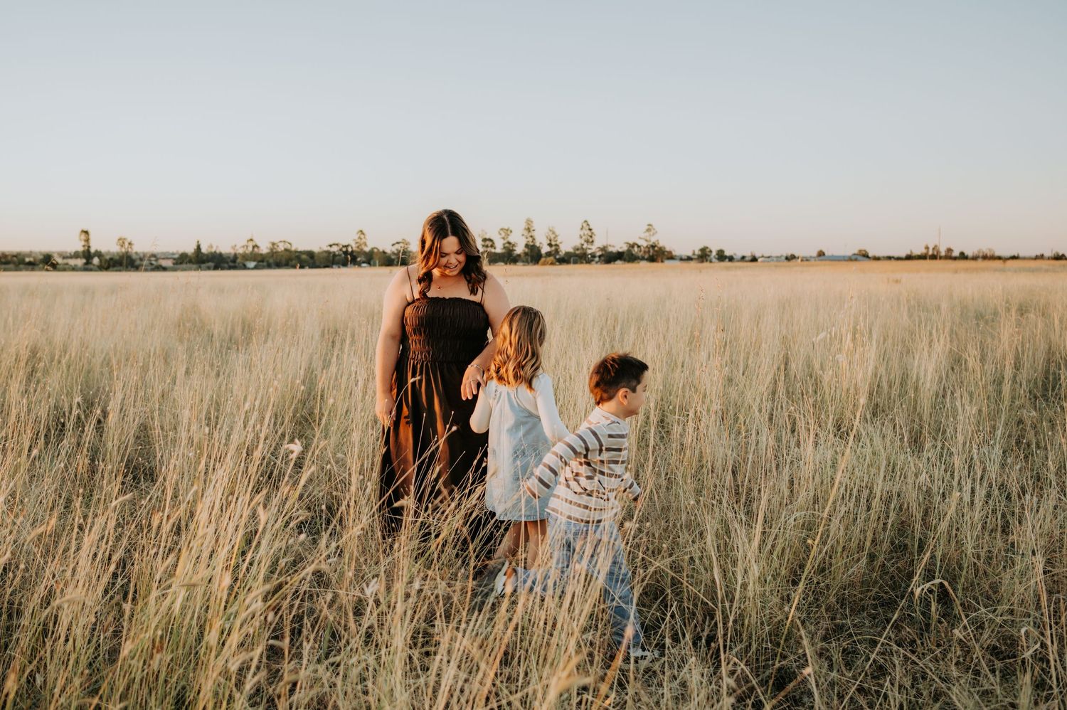 A woman and two children walk through tall golden grass in an open field at sunset.
