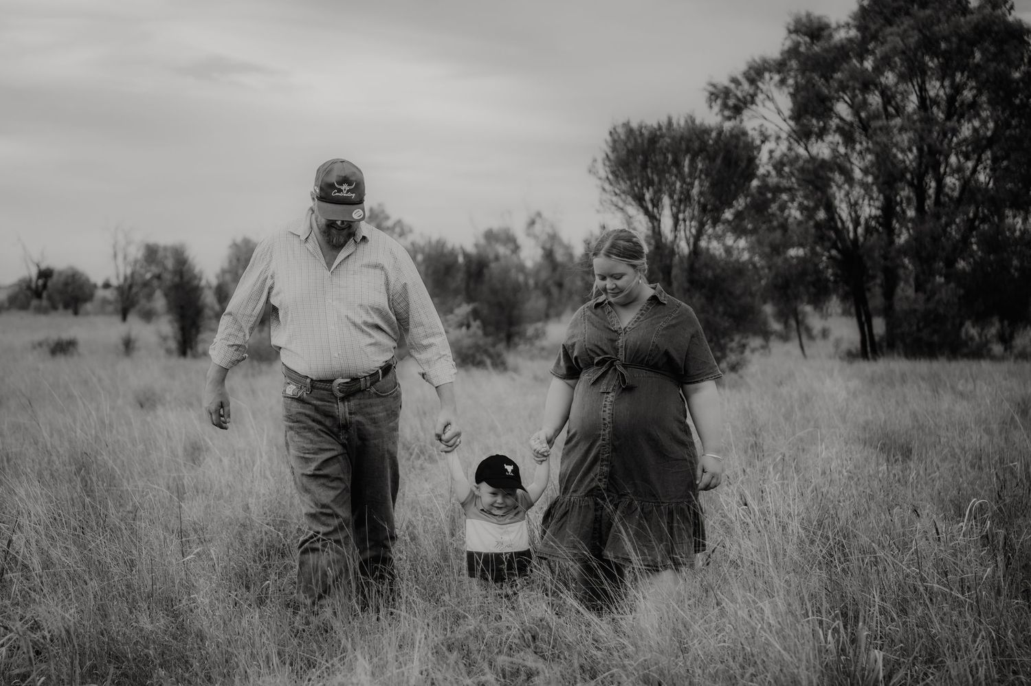 Black and white photo of a couple and toddler walking through a grassy field with trees in the background.