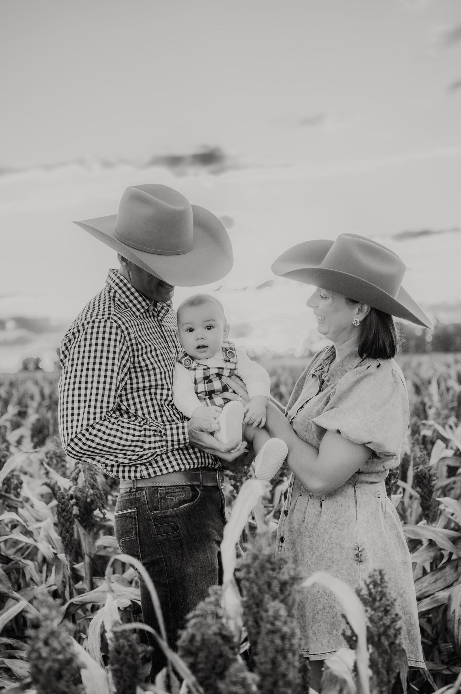 Black and white photo of a couple in cowboy hats holding their baby in a cornfield at sunset.
