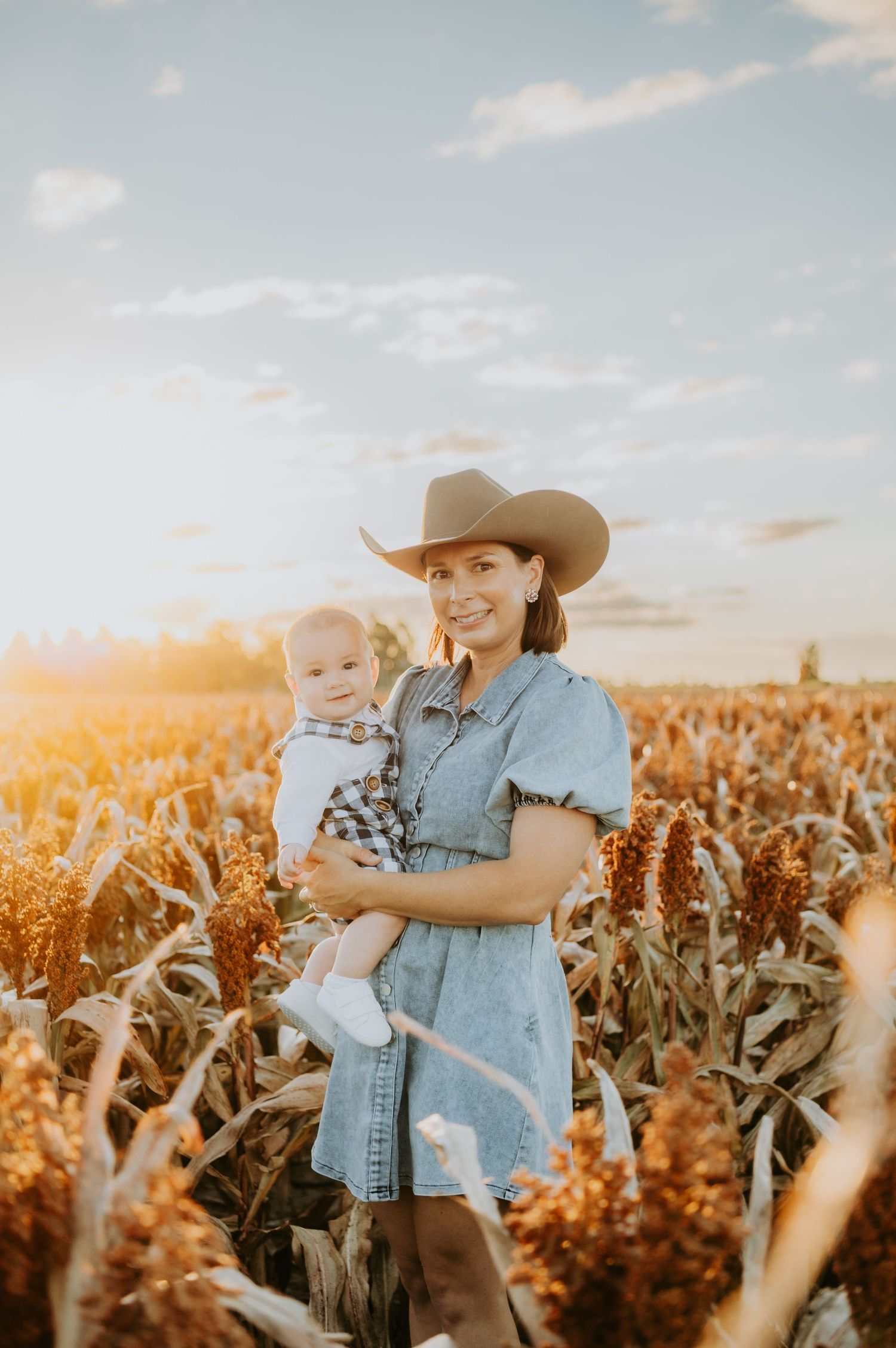 Woman in cowboy hat holding baby in golden sorghum field at sunset, wearing denim dress on a farm.