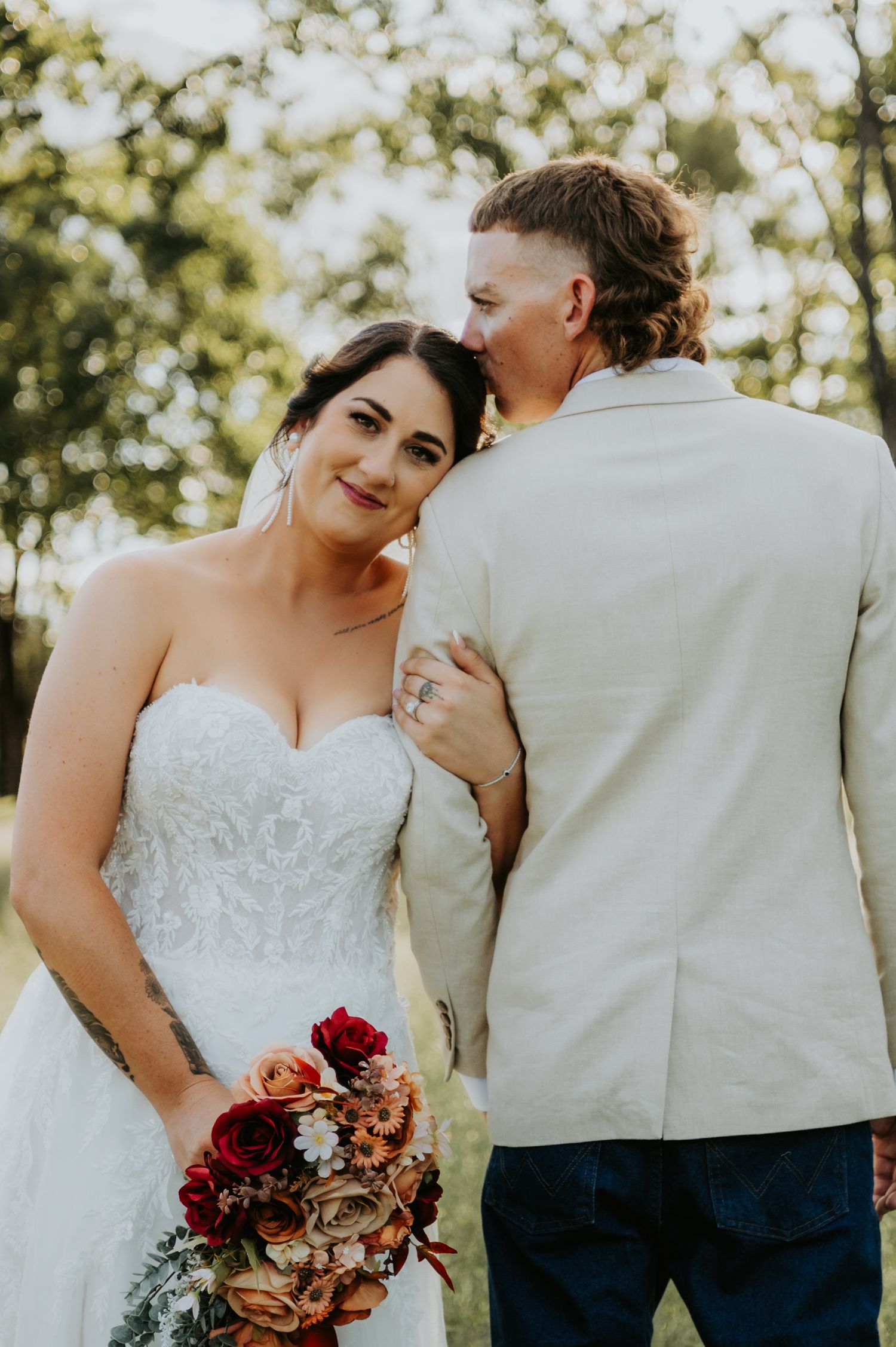 Bride in white lace gown holds red bouquet as groom in beige suit kisses her cheek outdoors.