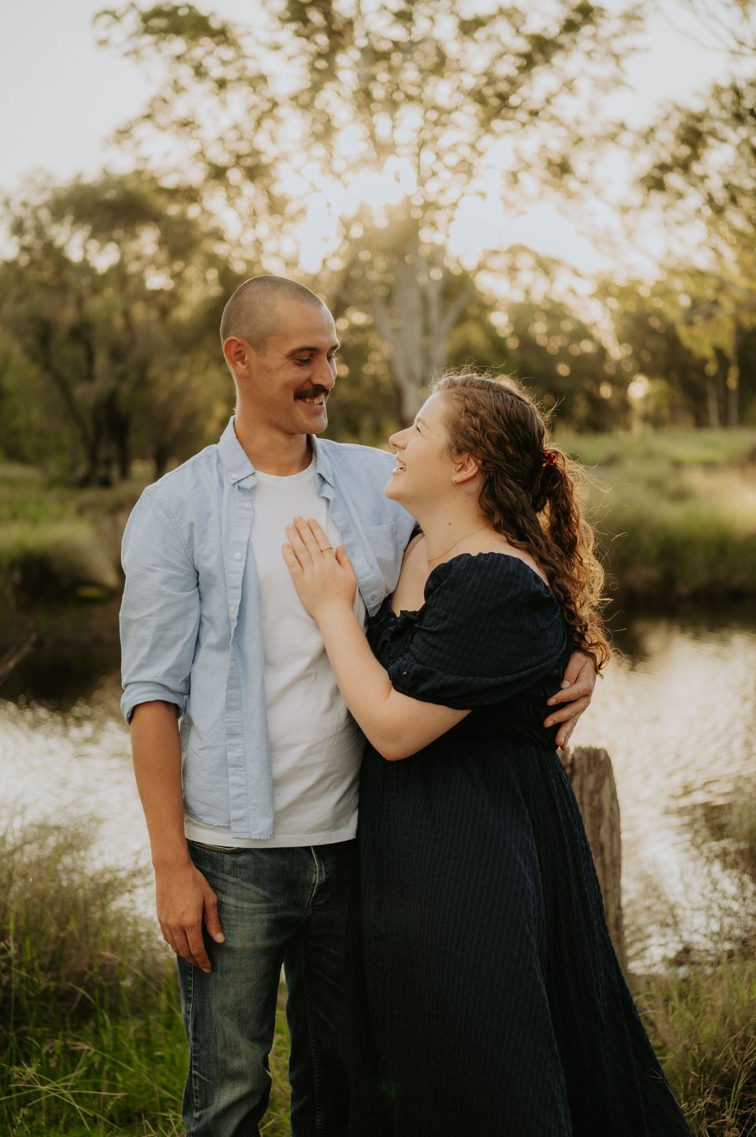 A couple sharing a tender moment outdoors near a pond, woman in black dress, man in blue shirt, golden hour light.