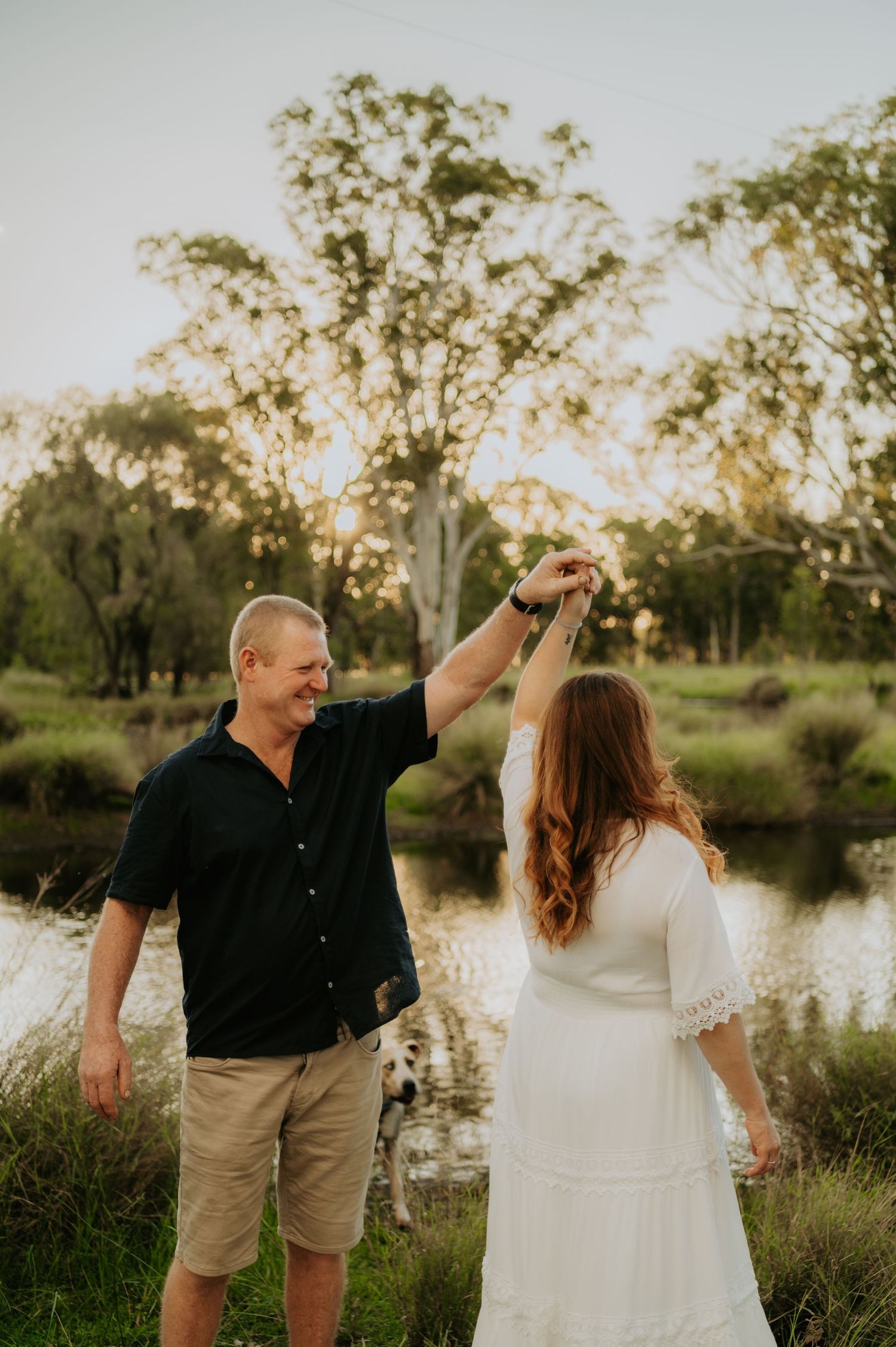A couple dances outdoors near a pond at golden hour, man in black shirt twirling woman in white dress.