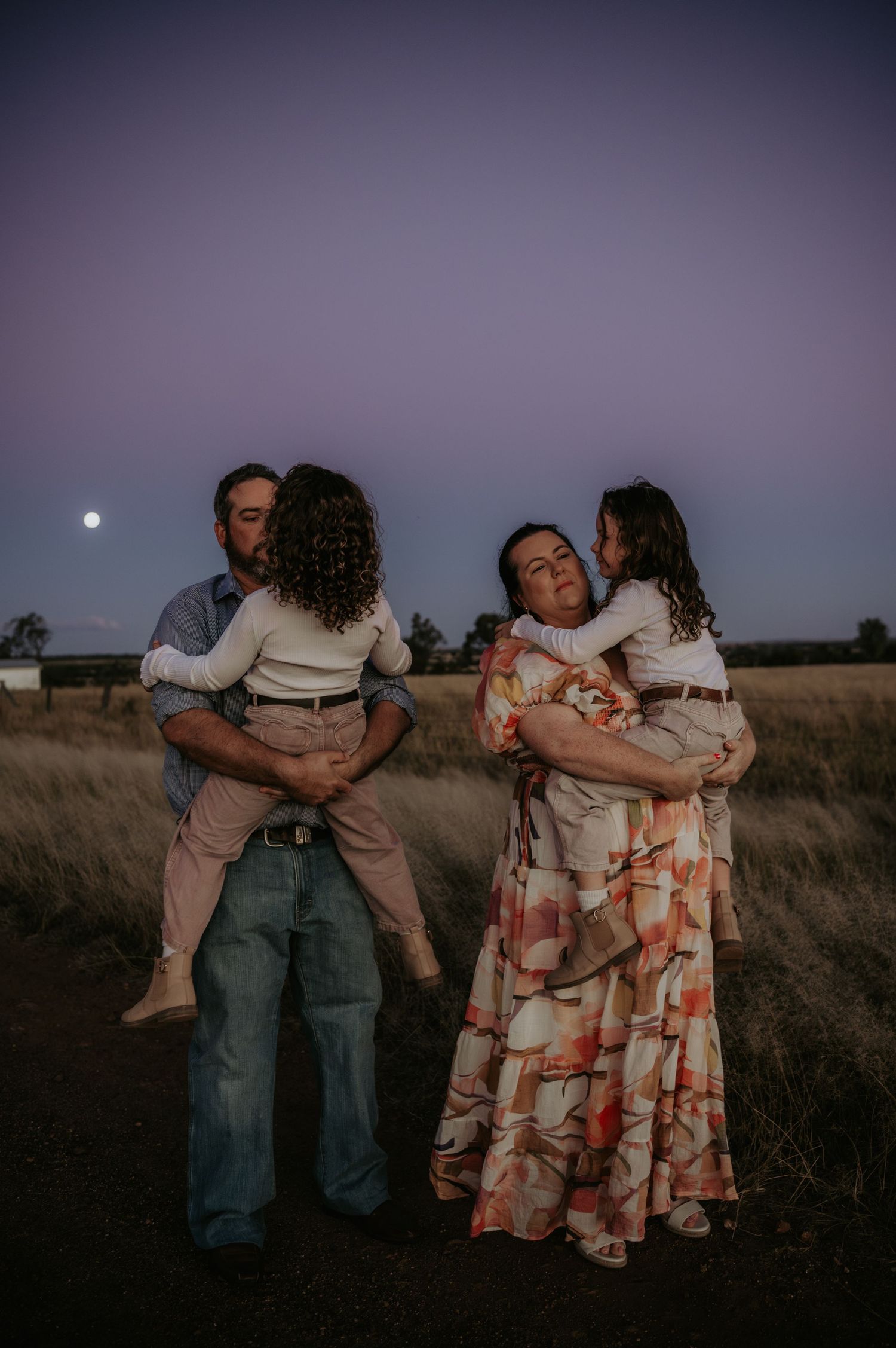 Family of four at dusk, parents holding children in a field with a full moon and purple sky.