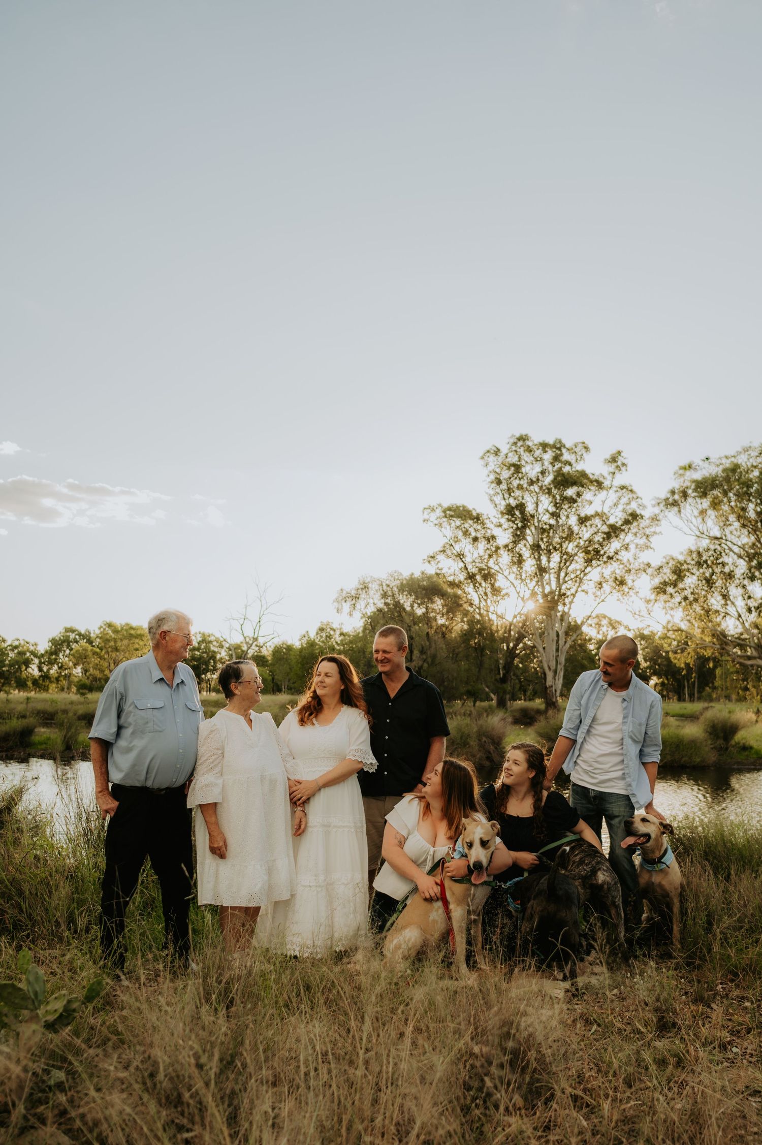 A joyful family group with two dogs poses in golden hour light among tall grass near a pond with eucalyptus trees.