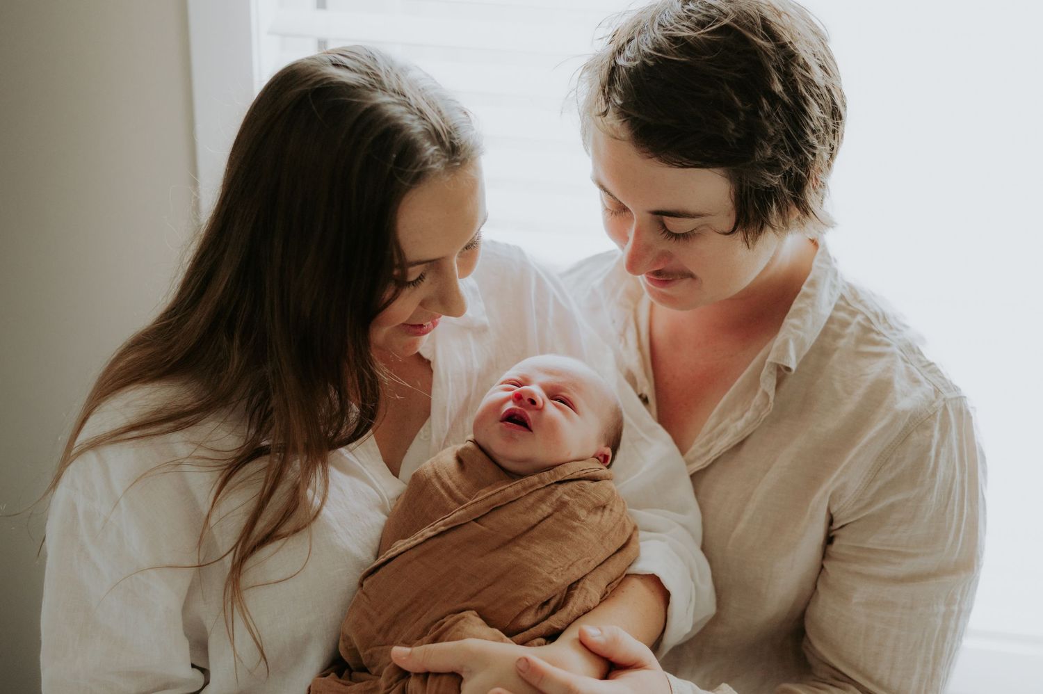 Two parents lovingly gaze at their swaddled newborn baby wrapped in a brown muslin cloth.