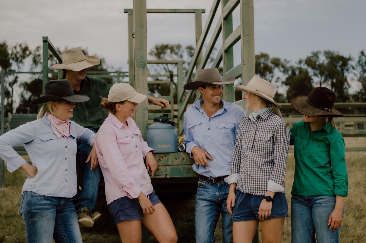 Group of five people in cowboy hats and jeans leaning against a farm trailer in a rural Australian setting.