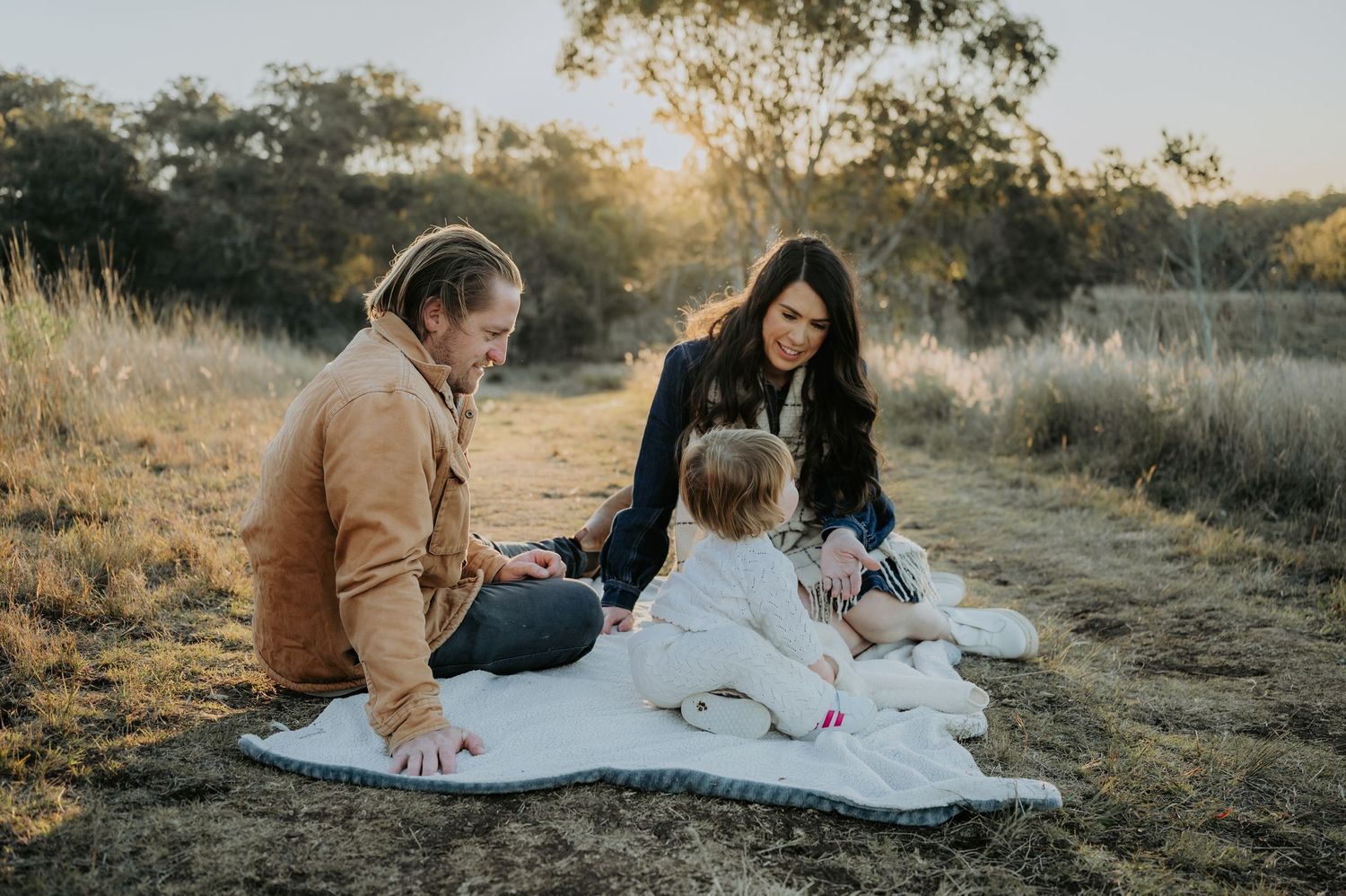 Family sitting on blanket in golden field at sunset, parents engaging with toddler in white outfit.