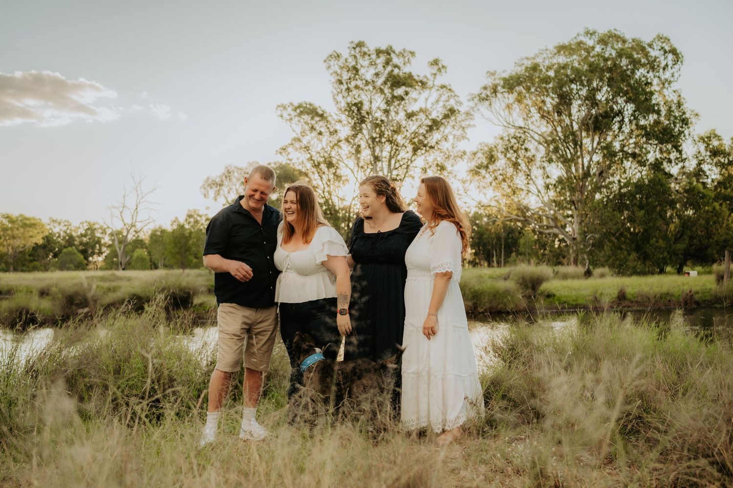 Family of four smiling together in a lush green field at golden hour, dressed in casual white and black outfits.