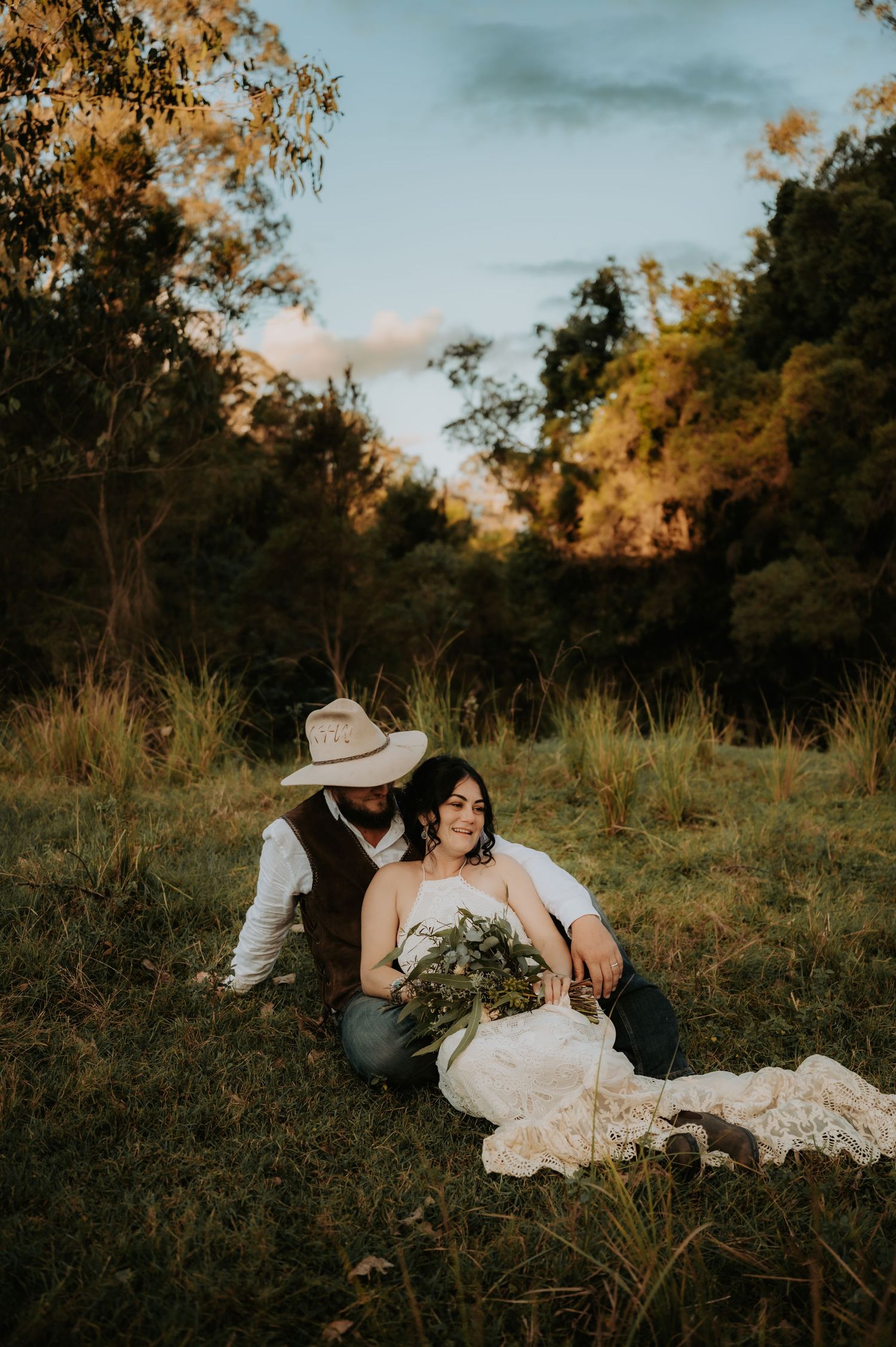 Bride and groom sitting together in a sunlit meadow, groom wearing a cowboy hat, bride holding a lush bouquet in wedding dress.