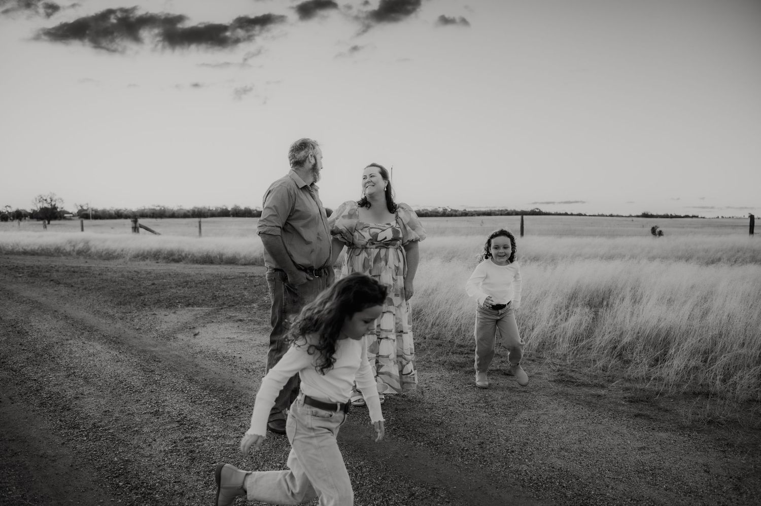 Black and white family photo on a rural dirt road at sunset, parents embracing while children play in a field.