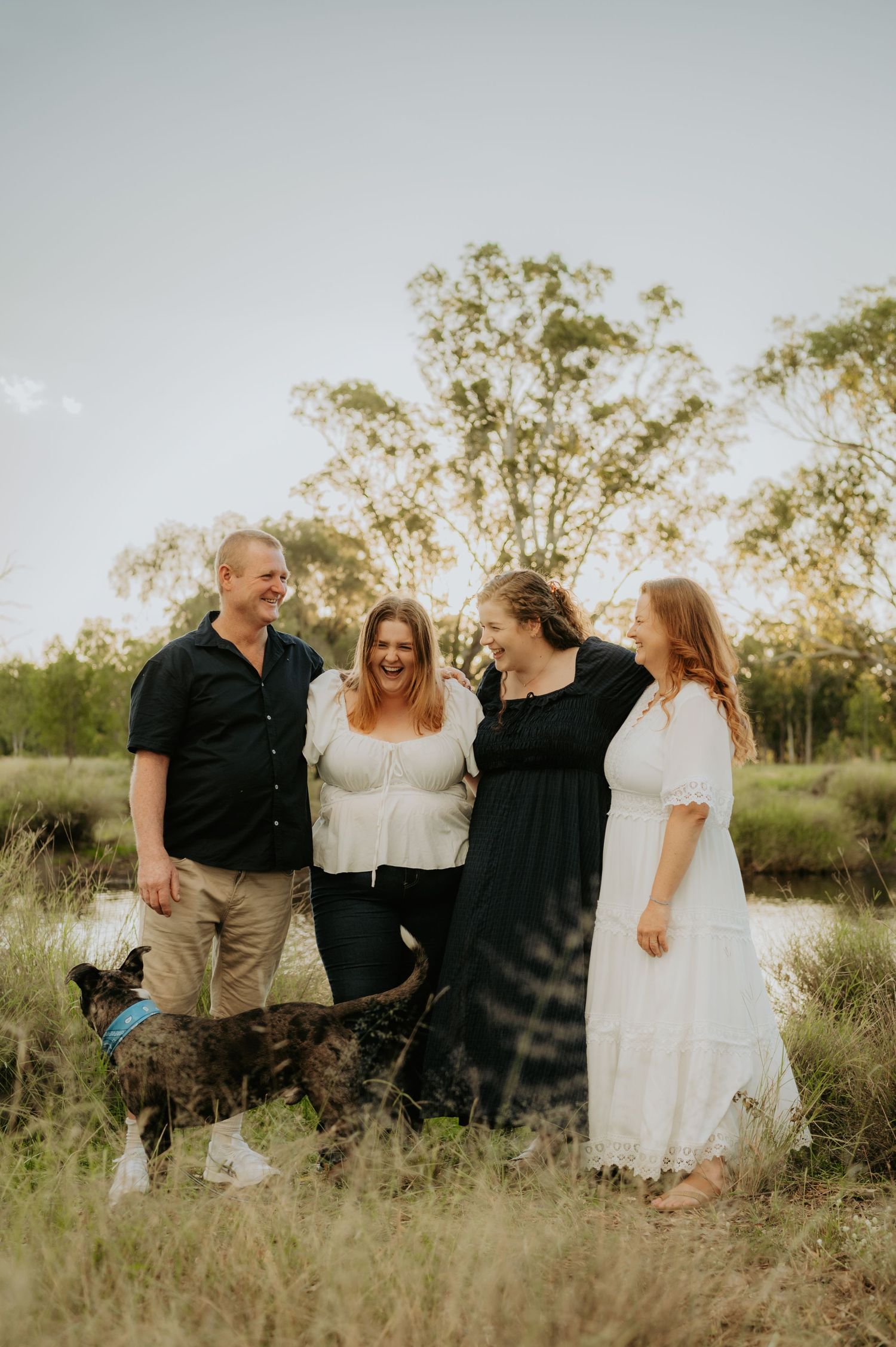 Family of four laughing together outdoors with their dog in a grassy field near a pond at golden hour.