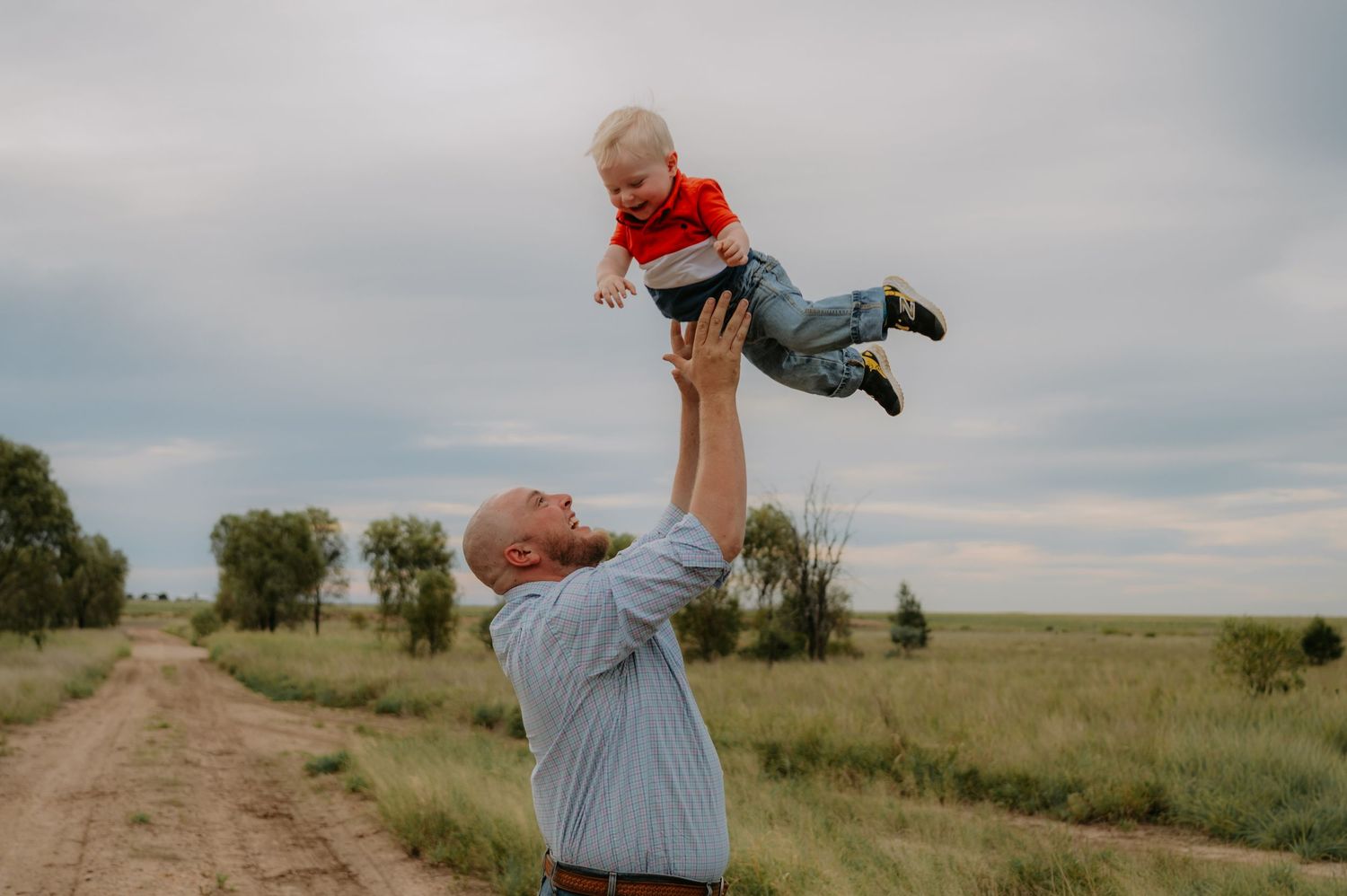 A person in a blue shirt playfully lifts a child into the air while standing in a grassy field at sunset.