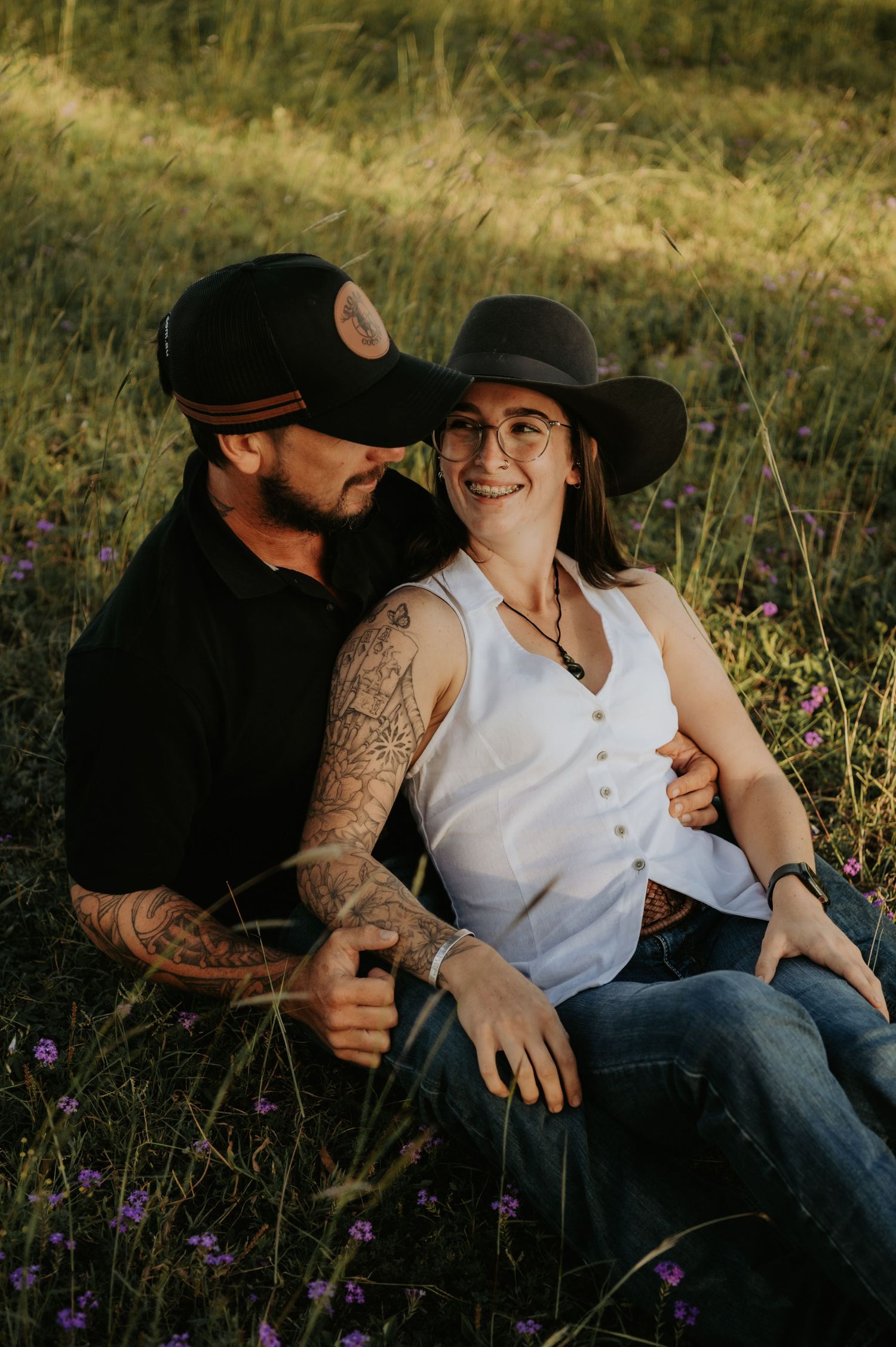 A couple sits together in a grassy field, the man with tattoos embracing a smiling woman in a wide-brim hat.