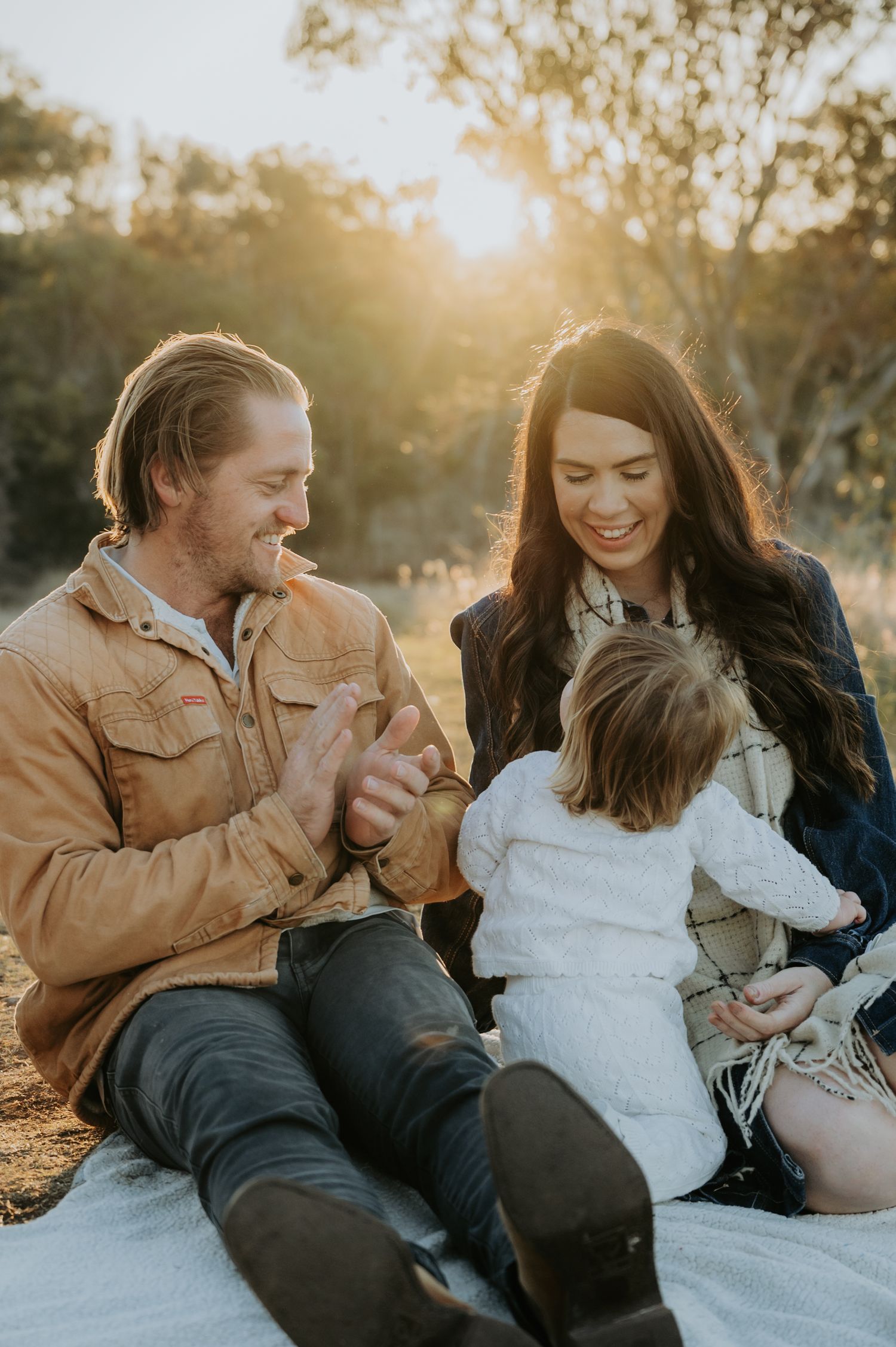 A happy family sitting outdoors at golden hour, parents smiling at their young child on a white blanket.