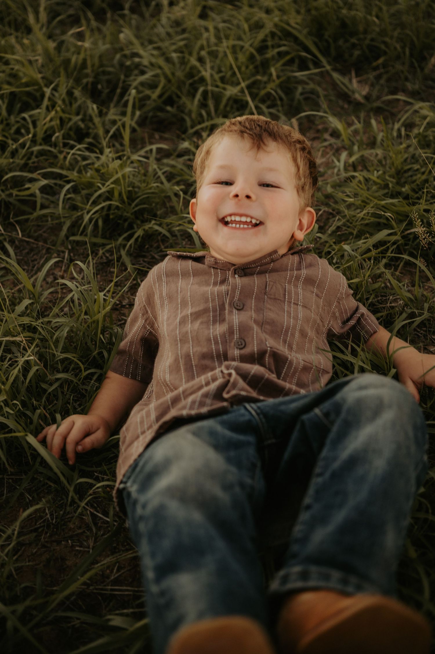 Child lying in grass with joyful expression wearing brown shirt and jeans.
