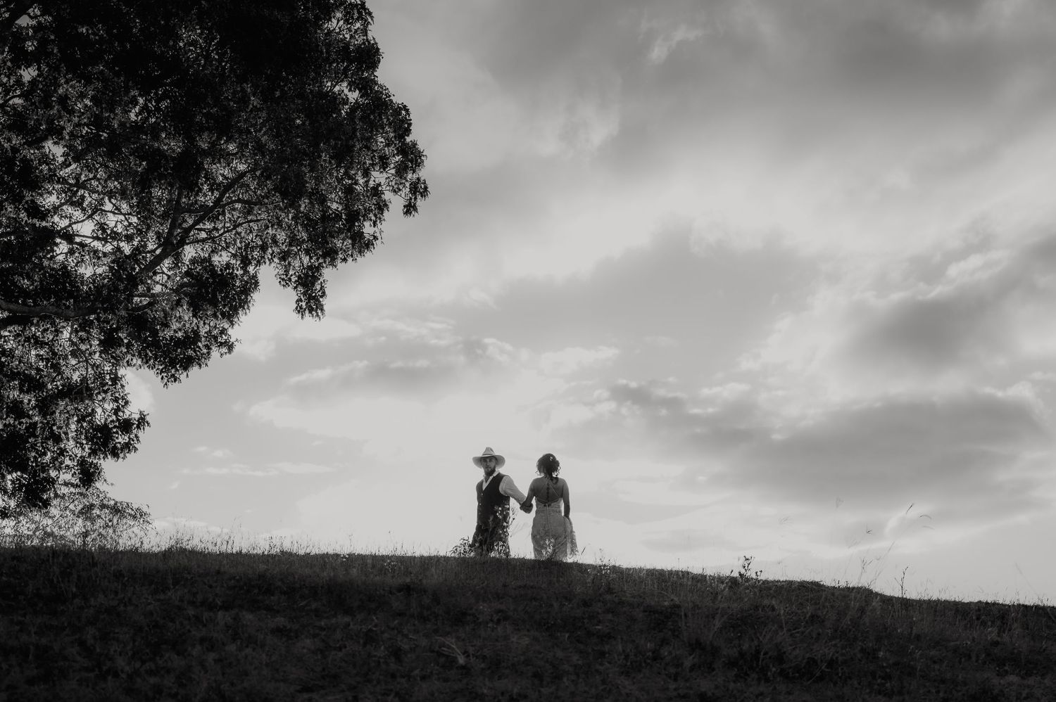 Black and white photo of two people in dresses standing on a hill near a tree under a cloudy sky.
