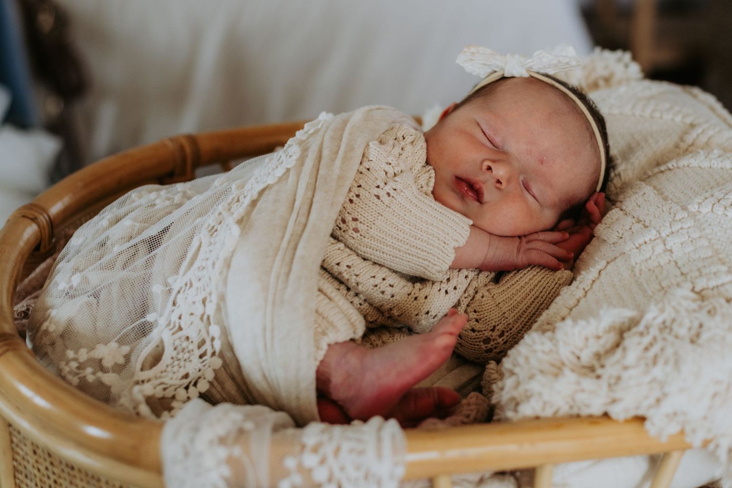 Peaceful newborn baby swaddled in cream wrap sleeping in a wicker bassinet with lace blankets.