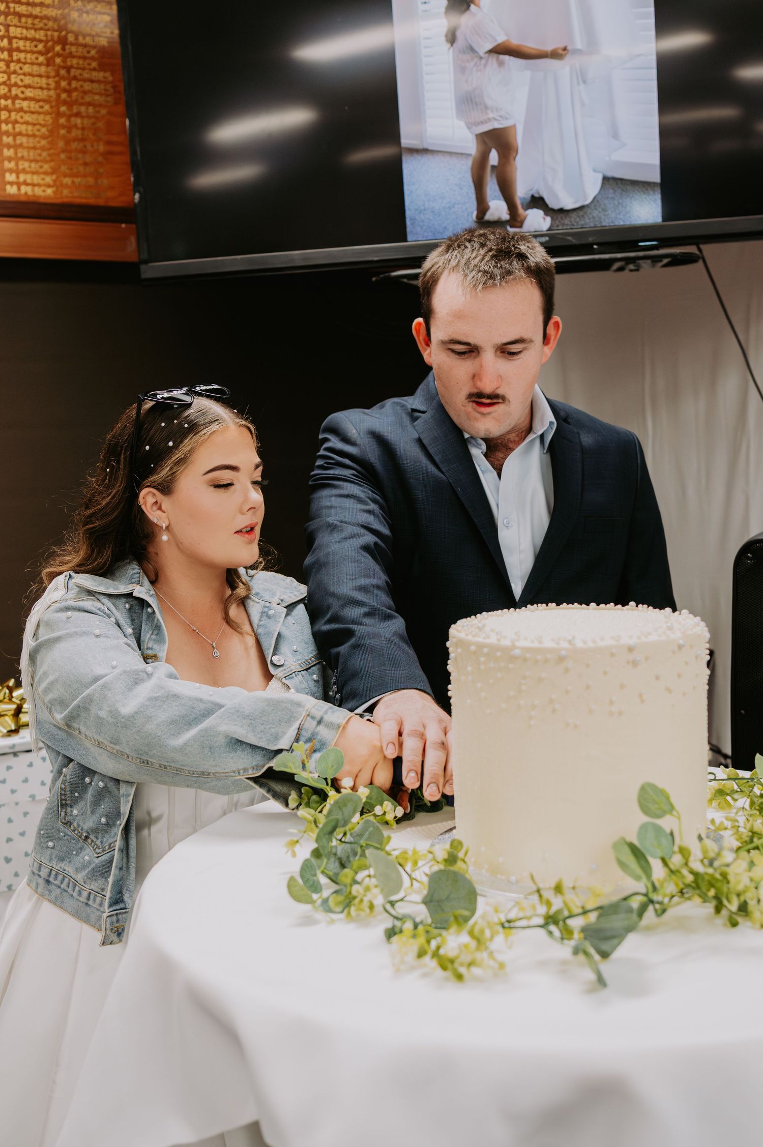 Bride and groom cutting a white wedding cake decorated with greenery at their reception venue.