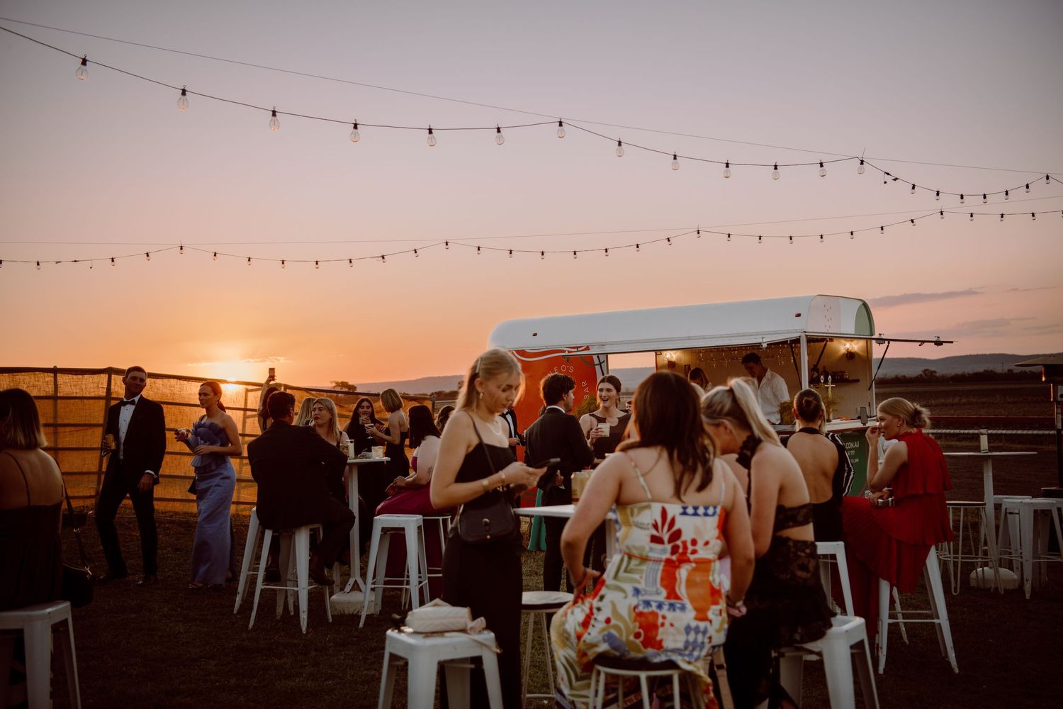 String lights illuminate an outdoor rooftop event space during golden hour as guests mingle and enjoy the sunset.