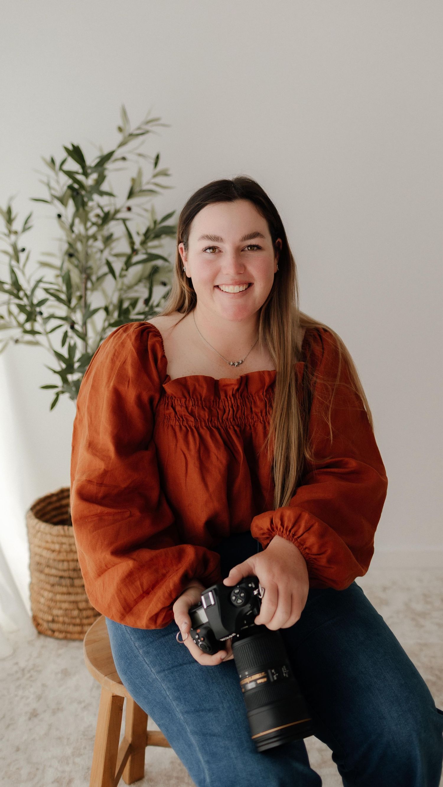 Photographer in orange blouse sits casually with professional camera in bright studio setting.