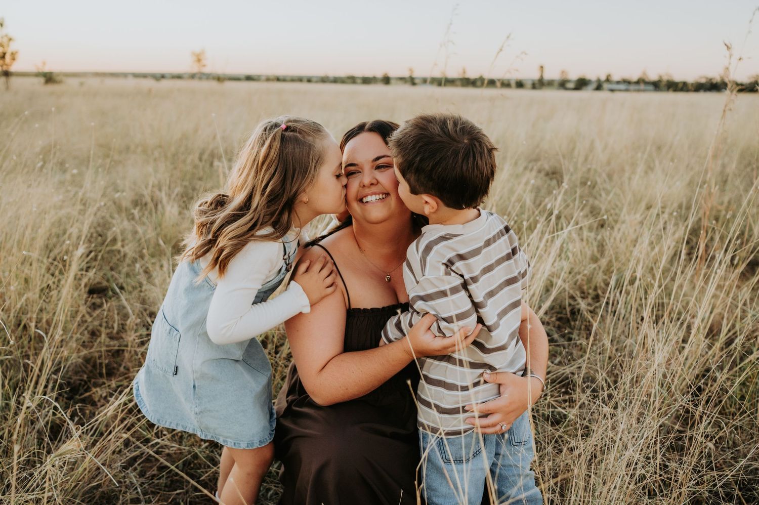 A mother smiles as her two young children kiss her cheeks in a golden grass field at sunset.
