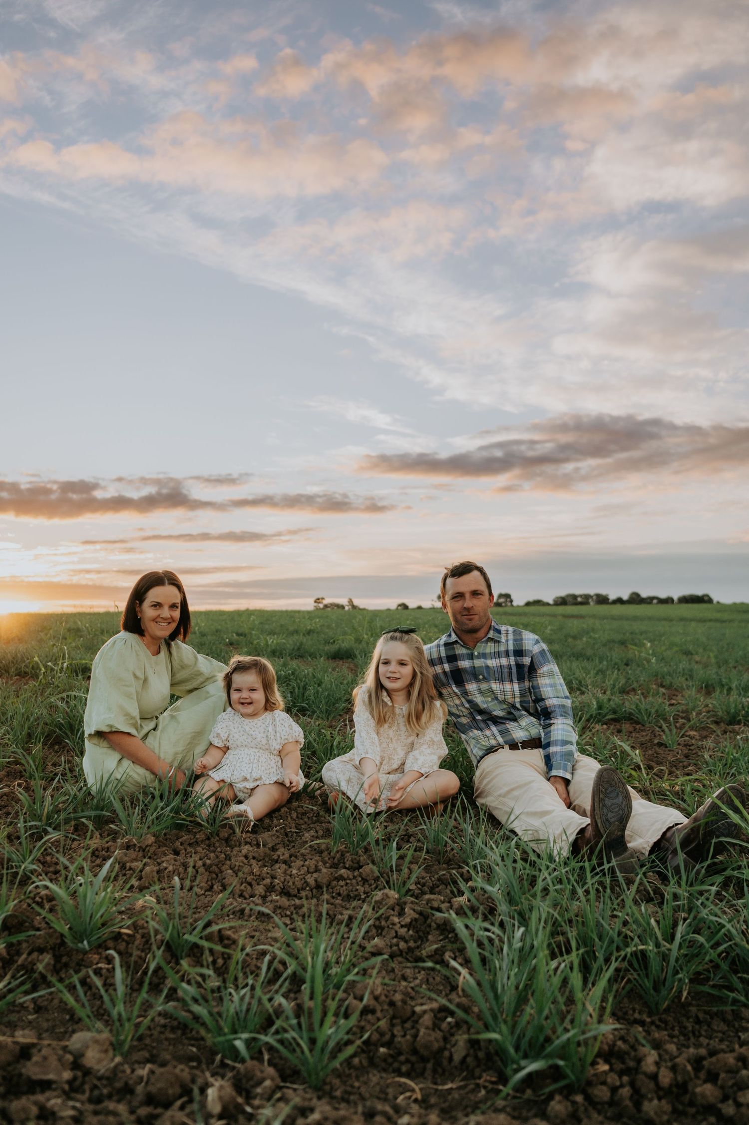 Family of four sitting in a green farm field at sunset, two young girls between their smiling parents.