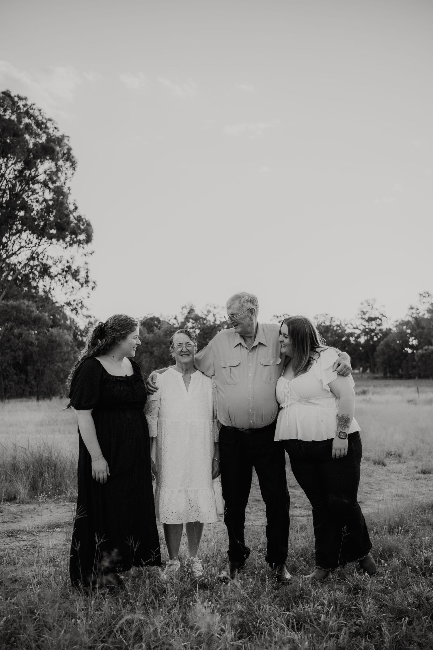 Black and white family portrait of four people laughing together in a rural field with trees in the background.